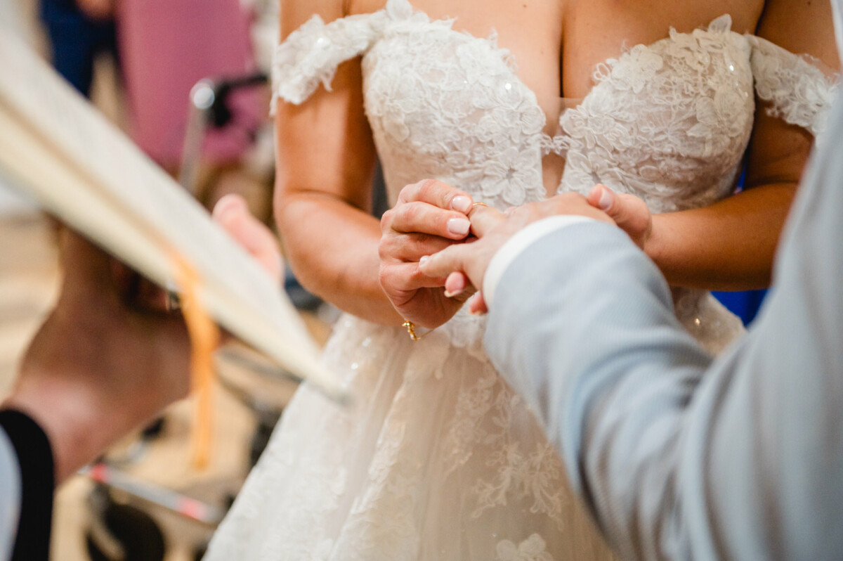 Eine Braut in einem weißen Spitzenkleid tauscht während einer Hochzeitszeremonie die Ringe mit einem Bräutigam in einem hellen Anzug.