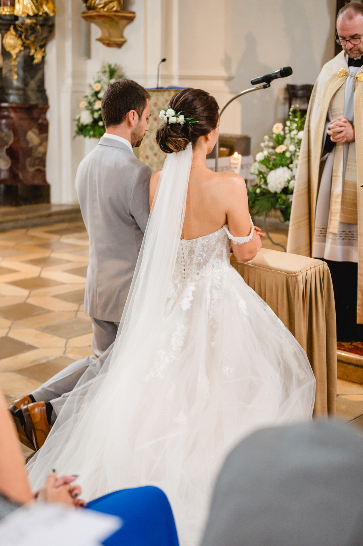 Braut und Bräutigam knien während einer Hochzeitszeremonie am Altar, während ein Priester in der Nähe steht. Die Braut trägt ein schulterfreies Kleid und einen Schleier; der Bräutigam trägt einen hellgrauen Anzug.