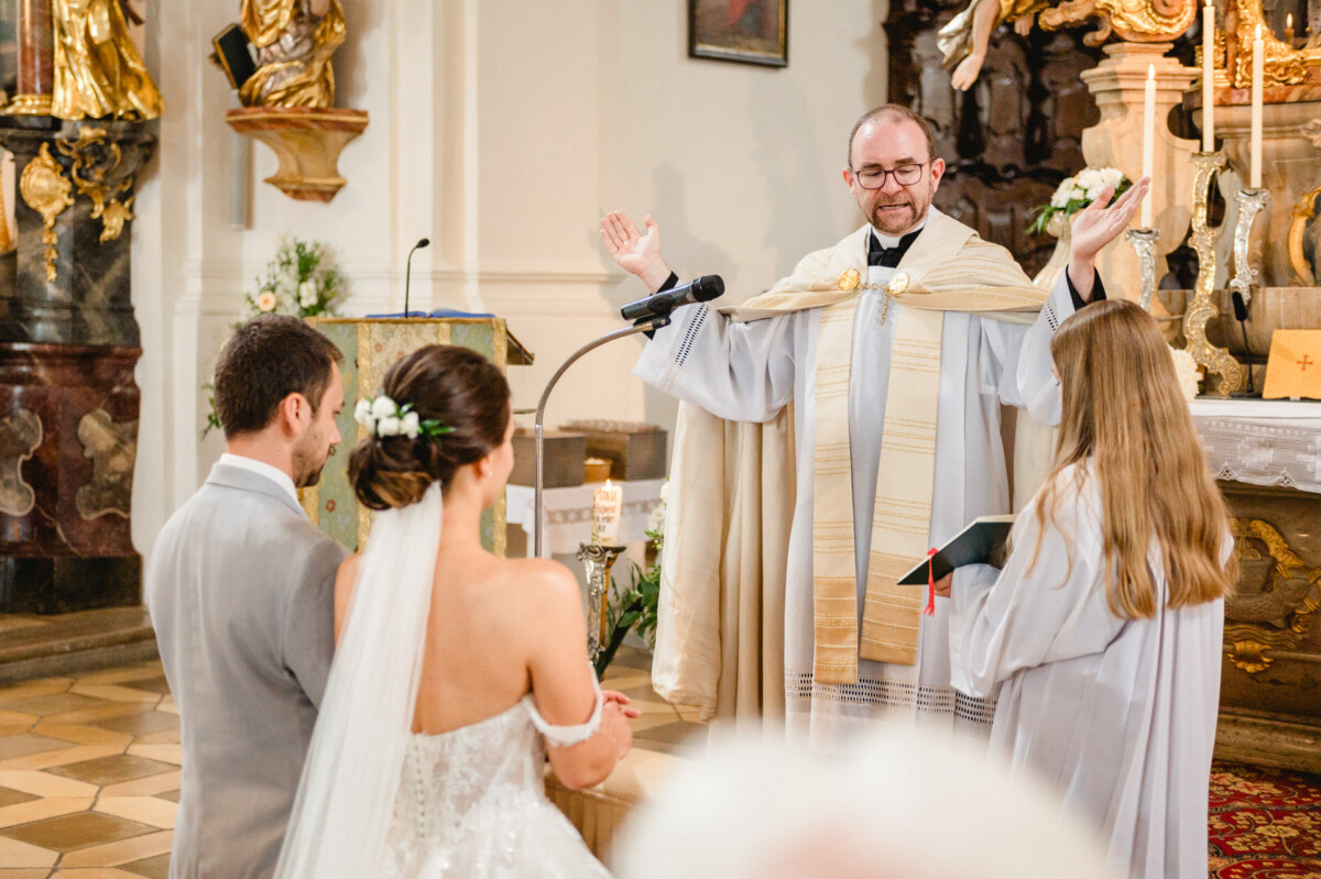 Ein Priester führt in einer Kirche eine Hochzeitszeremonie durch, während Braut und Bräutigam ihm gegenübersitzen.
