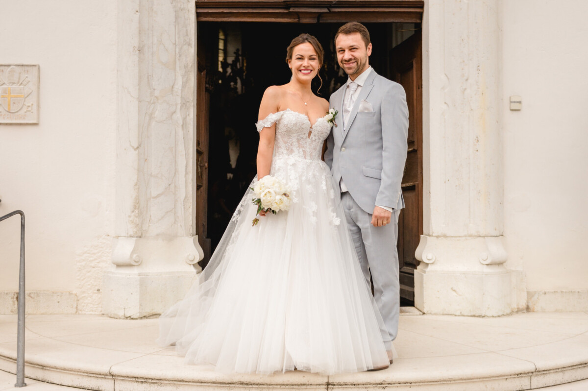 Eine Braut im weißen Kleid und ein Bräutigam im hellen Anzug stehen lächelnd in einer Tür. Die Braut hält einen Strauß weißer Blumen in der Hand.