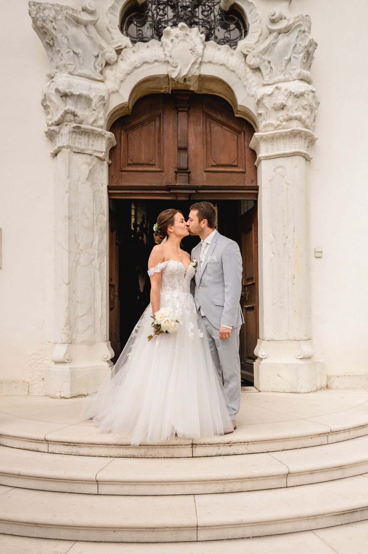 Braut und Bräutigam küssen sich vor dem Eingang eines dekorativen Gebäudes. Die Braut hält einen Blumenstrauß und beide sind in Abendgarderobe gekleidet.
