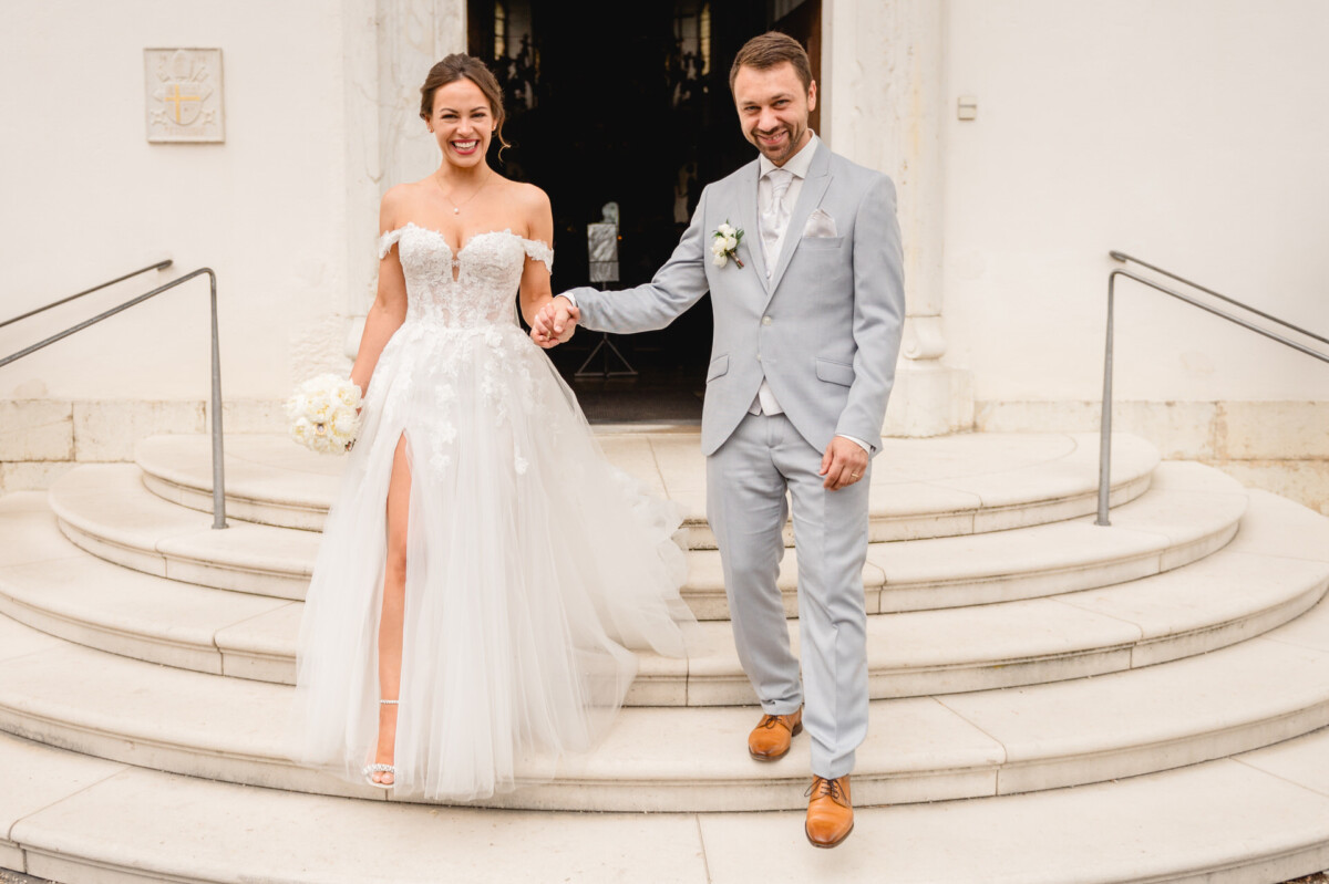 Ein Paar im Hochzeitskleid geht Händchen haltend Marmorstufen hinab. Die Braut trägt ein schulterfreies weißes Kleid mit einem Blumenstrauß und der Bräutigam einen hellgrauen Anzug mit braunen Schuhen.