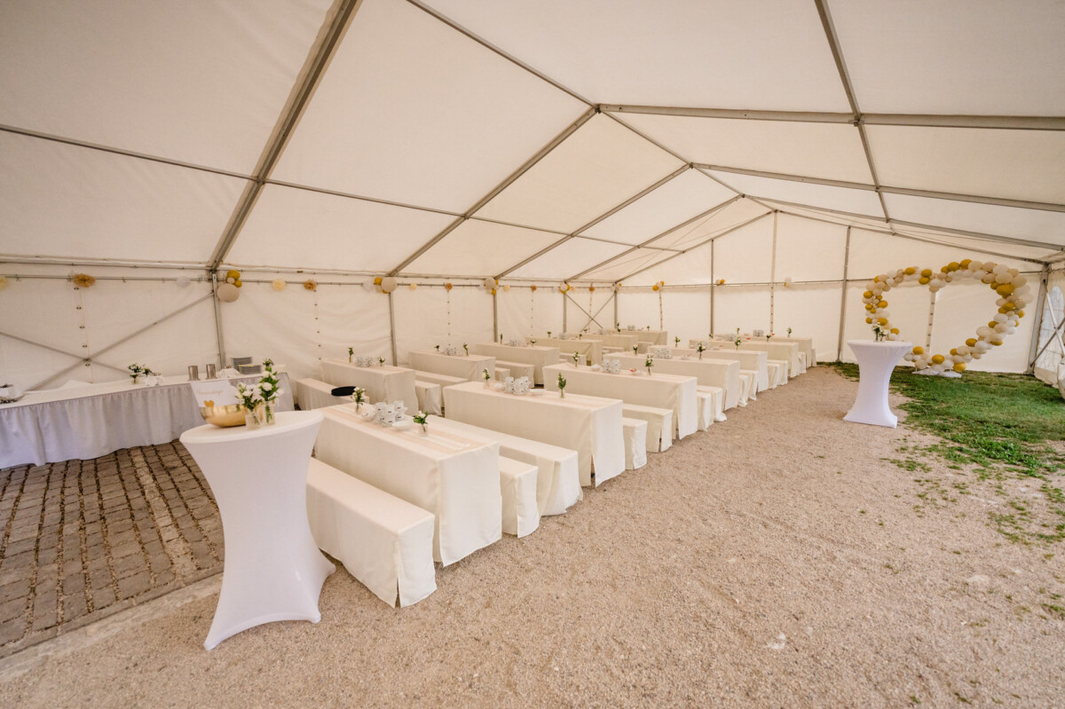 Weißes Partyzelt mit langen Tischen und Bänken, bedeckt mit weißem Stoff, dekoriert mit Blumen und einem Ballonbogen auf der rechten Seite.