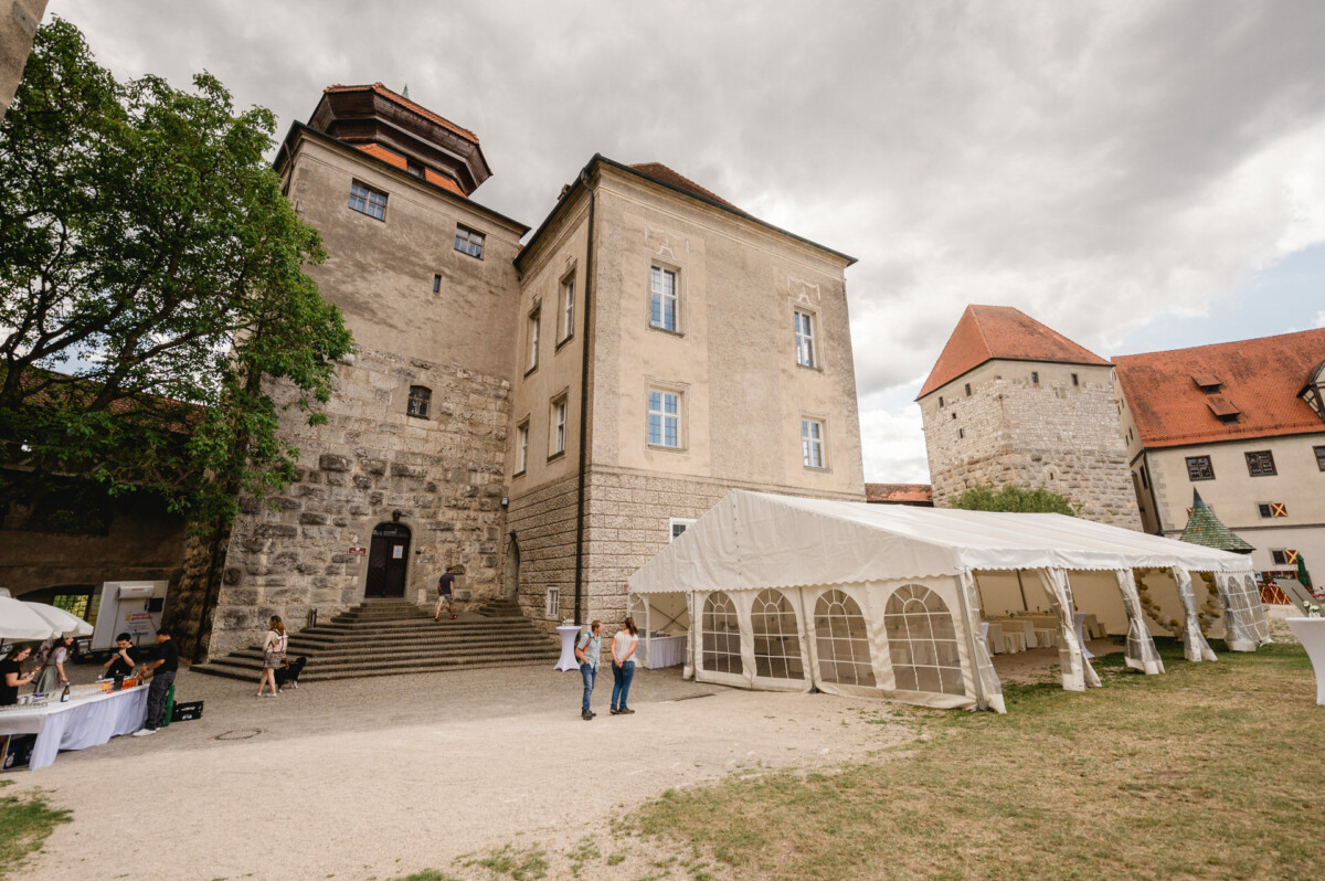 Ein Steingebäude mit einem hohen Turm und einem weißen Zelt daneben. Zwei Personen stehen daneben und links stehen Tische mit Sonnenschirmen. Über uns ein bewölkter Himmel.