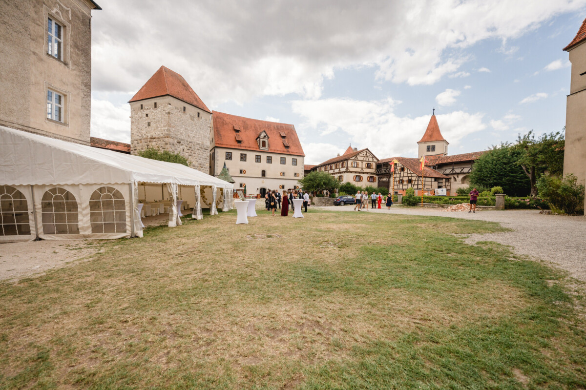 Ein Hochzeitsempfang im Innenhof eines historischen Gebäudes, mit geselligen Menschen, links einem weißen Zelt und im Hintergrund einem geparkten Auto unter einem bewölkten Himmel.