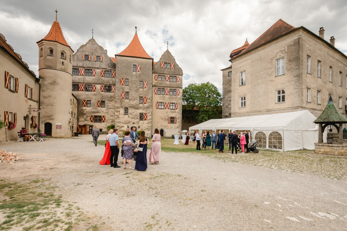 An einem bewölkten Tag versammeln sich Menschen vor einem alten Schloss mit roten Dächern und einem weißen Veranstaltungszelt.