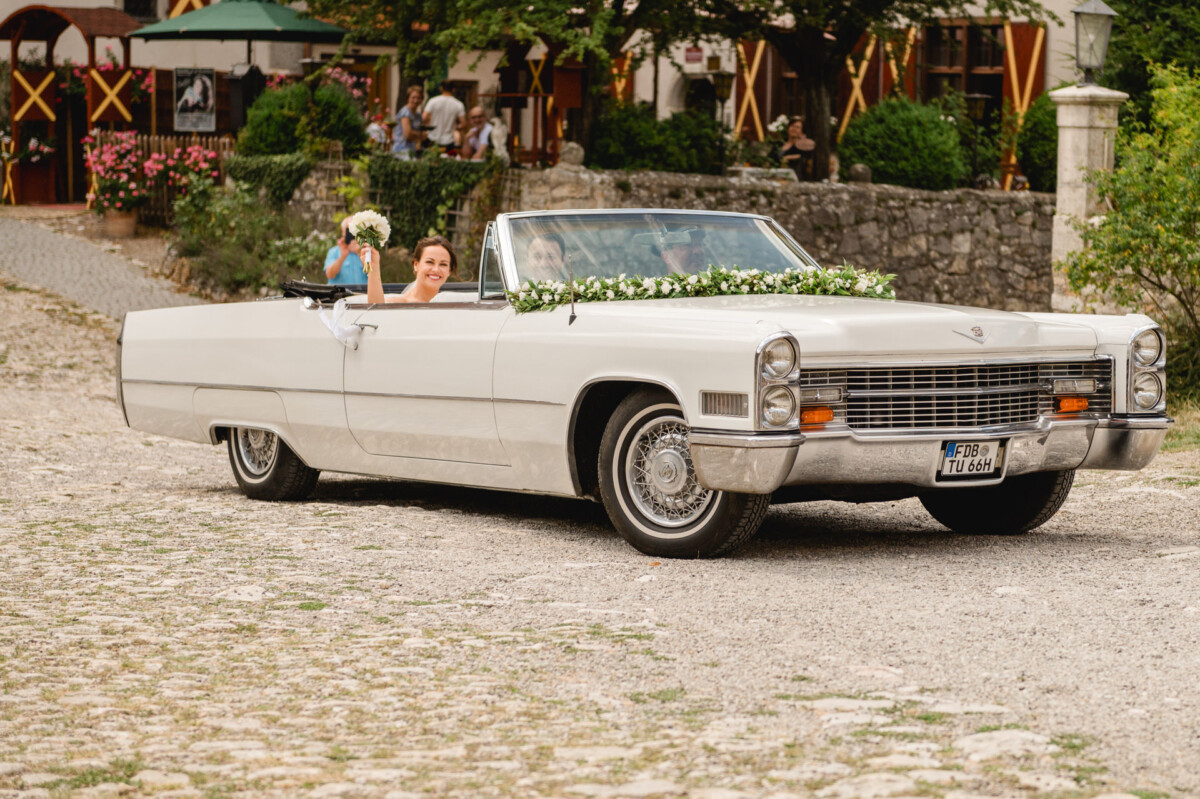Braut und Bräutigam fahren in einem mit Blumen geschmückten weißen Oldtimer-Cabrio und winken, während sie eine Kopfsteinpflasterstraße entlangfahren.