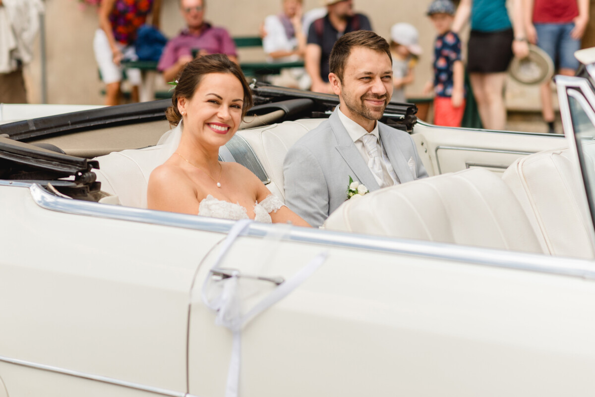 Braut und Bräutigam lächeln, während sie in einem weißen Cabrio sitzen, im Hintergrund sind Zuschauer zu sehen.