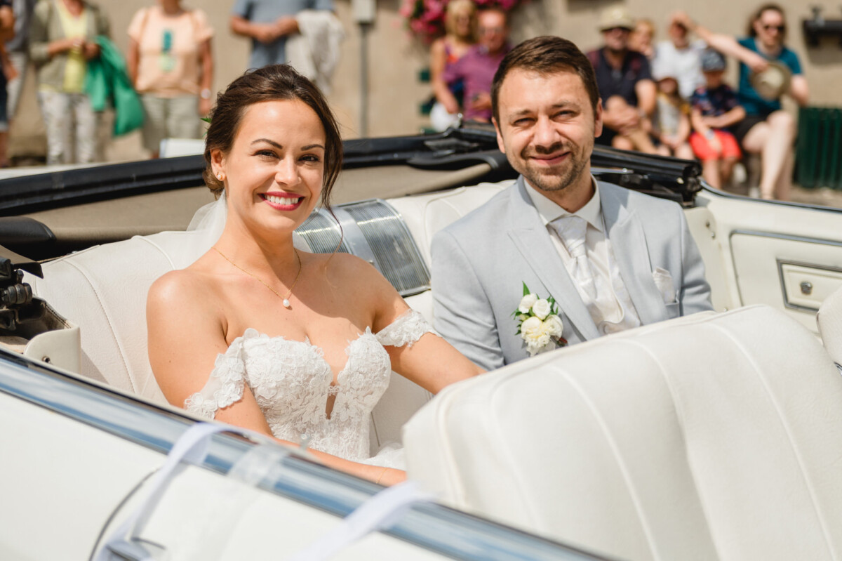 Braut und Bräutigam sitzen lächelnd in einem weißen Cabrio mit offenem Verdeck. Die Braut trägt ein weißes Spitzenkleid und der Bräutigam einen hellgrauen Anzug mit einer Ansteckblume.