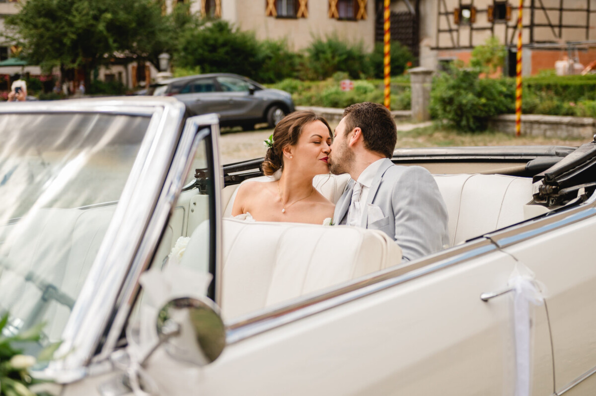 Ein Paar im Hochzeitskleid küsst sich auf dem Rücksitz eines weißen Cabrios.