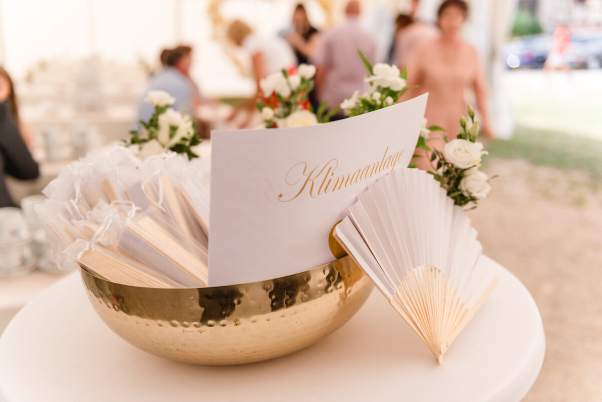In einer Messingschale liegen Hochzeitsprogramme und Faltfächer, um die sich kleine weiße Blumen tummeln. Im Hintergrund sind verschwommen Menschen zu sehen.