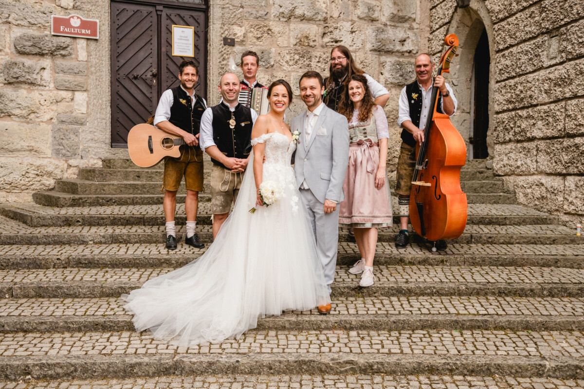Braut und Bräutigam posieren auf Steinstufen mit einer Band von Musikern in traditioneller Kleidung, darunter Gitarren und ein Kontrabass, vor der Kulisse eines alten Steingebäudes.