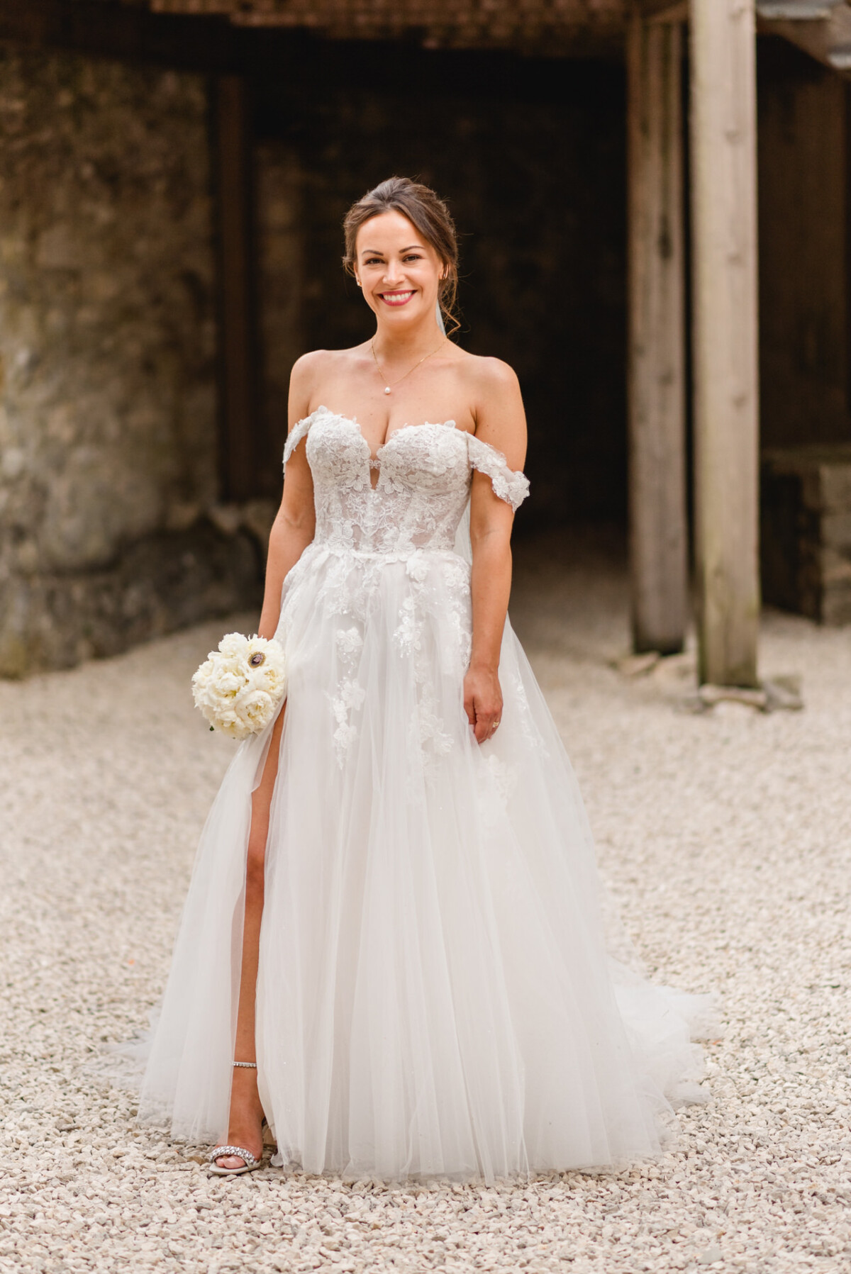 Eine Frau in einem schulterfreien Spitzenbrautkleid steht auf einem Kiesweg und hält einen Strauß weißer Blumen in der Hand. Im Hintergrund ist ein Steingebäude zu sehen.