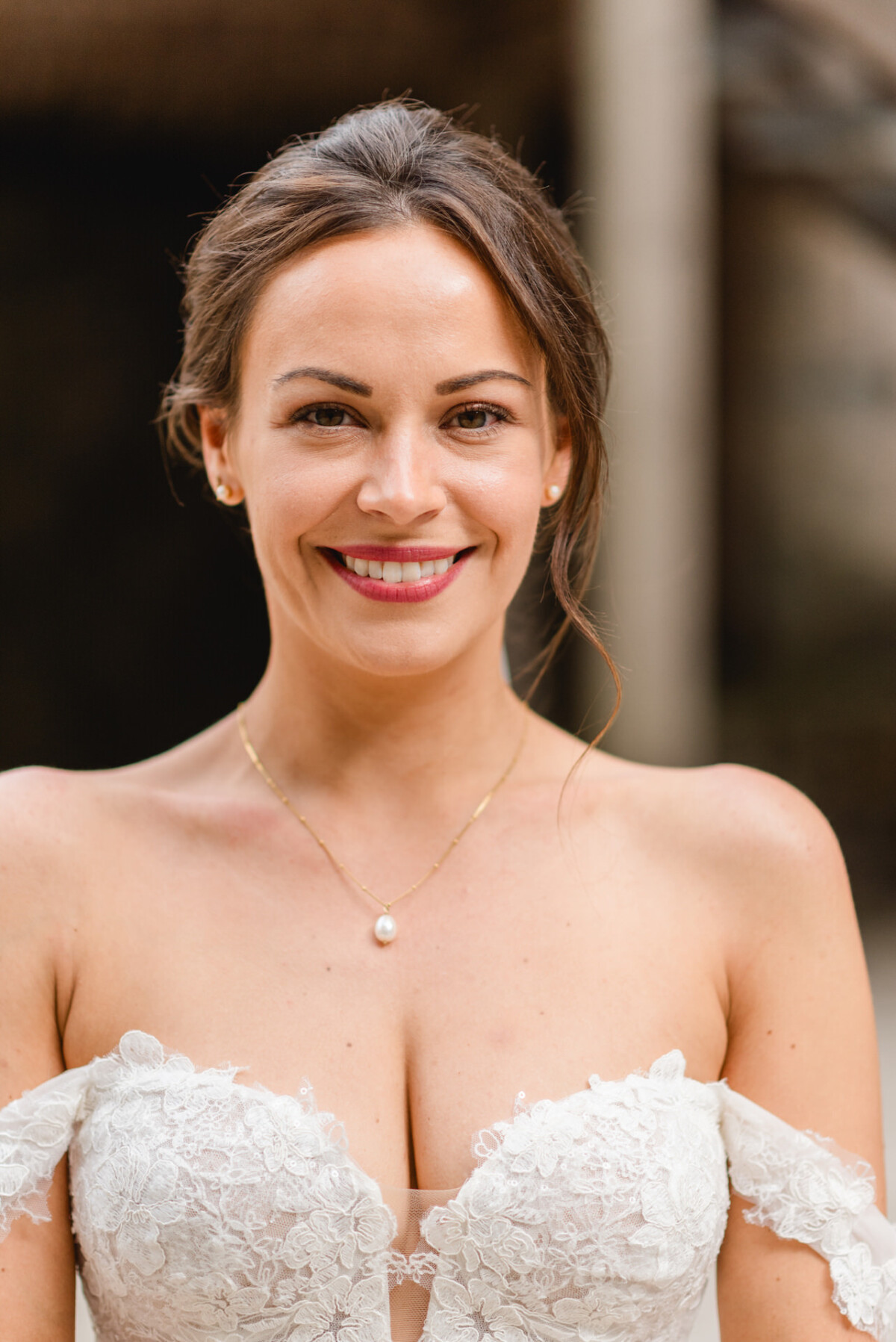 Lächelnde Frau in einem Spitzenbrautkleid mit schulterfreien Ärmeln und einer Perlenkette.