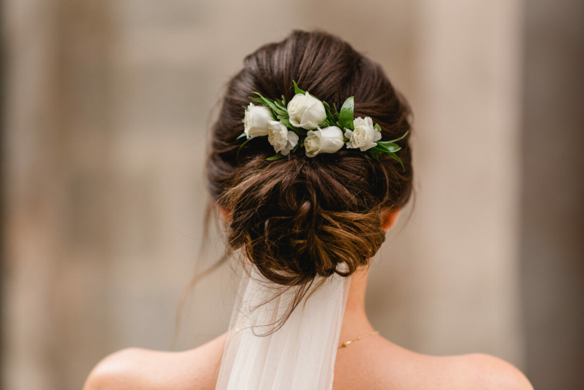 Eine Frau mit dunklem Haar, das zu einem tiefen Dutt frisiert ist, mit weißen Rosen und einem Schleier geschmückt ist, von hinten gesehen.