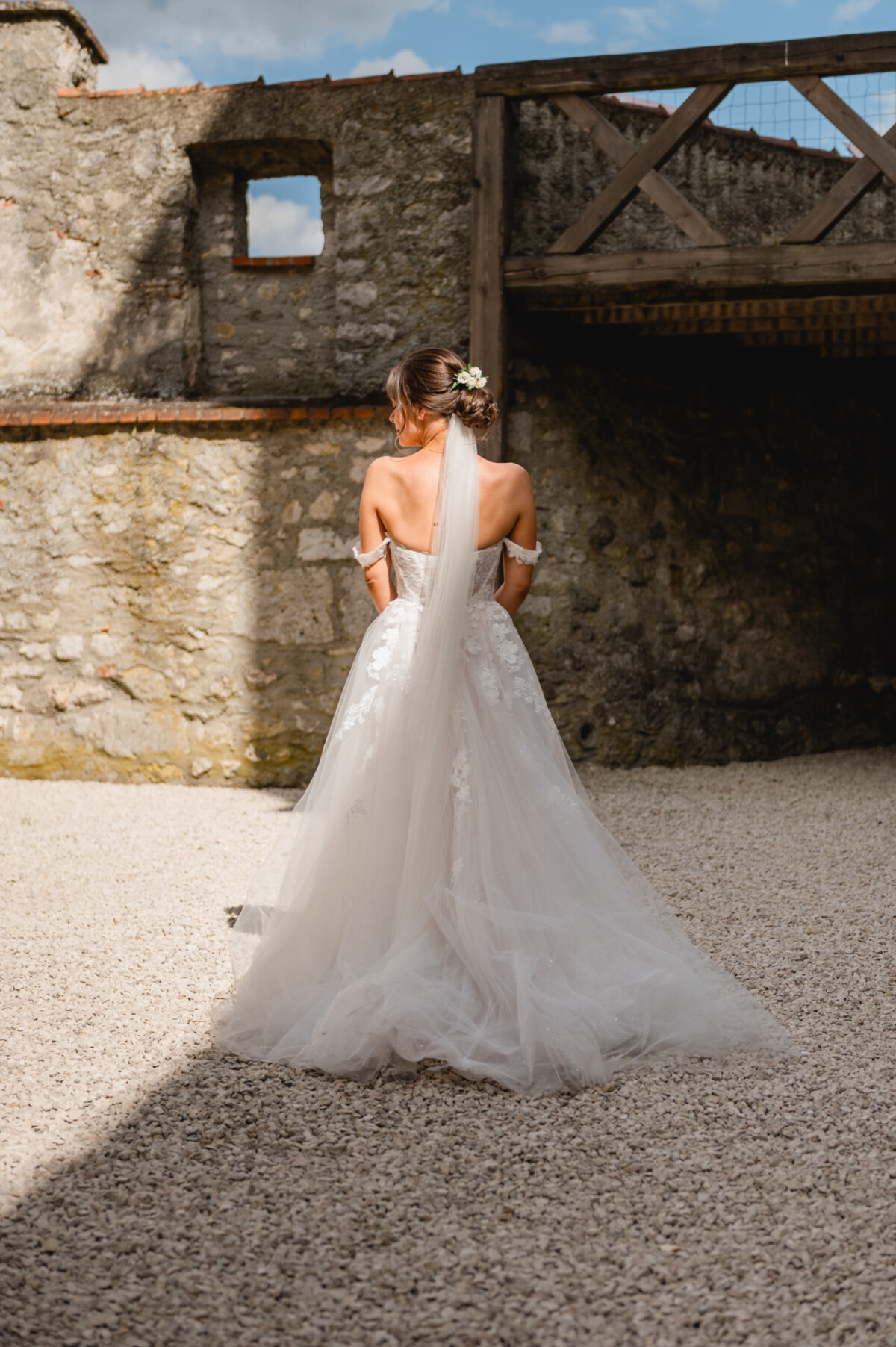 Eine Braut in einem weißen Kleid und einem langen Schleier steht an einem sonnigen Tag auf Kies vor einer Steinmauer und blickt abgewandt.