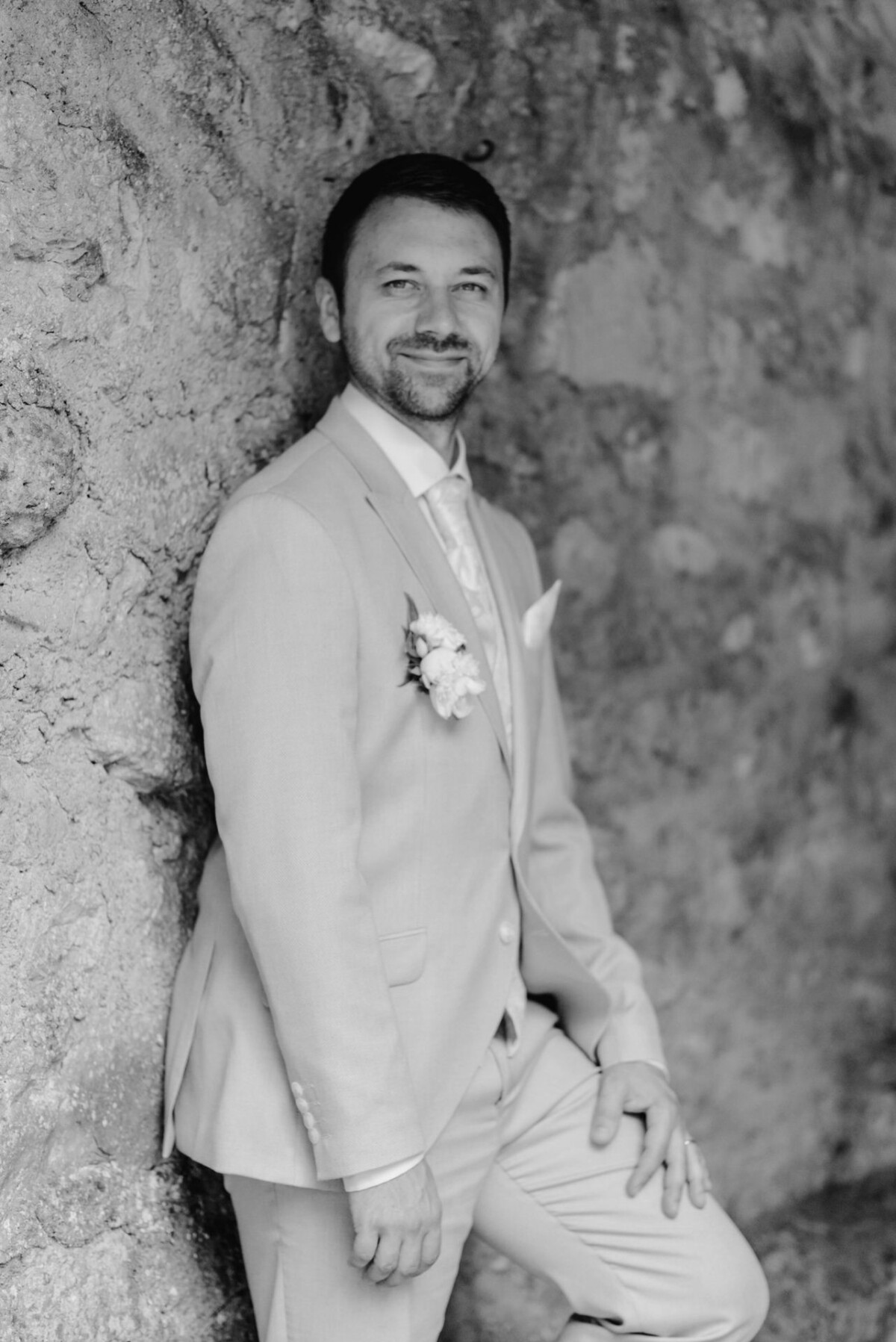 Ein Mann im Anzug mit einer Boutonniere steht vor einer strukturierten Wand, blickt in die Kamera und lächelt. Das Bild ist schwarzweiß.