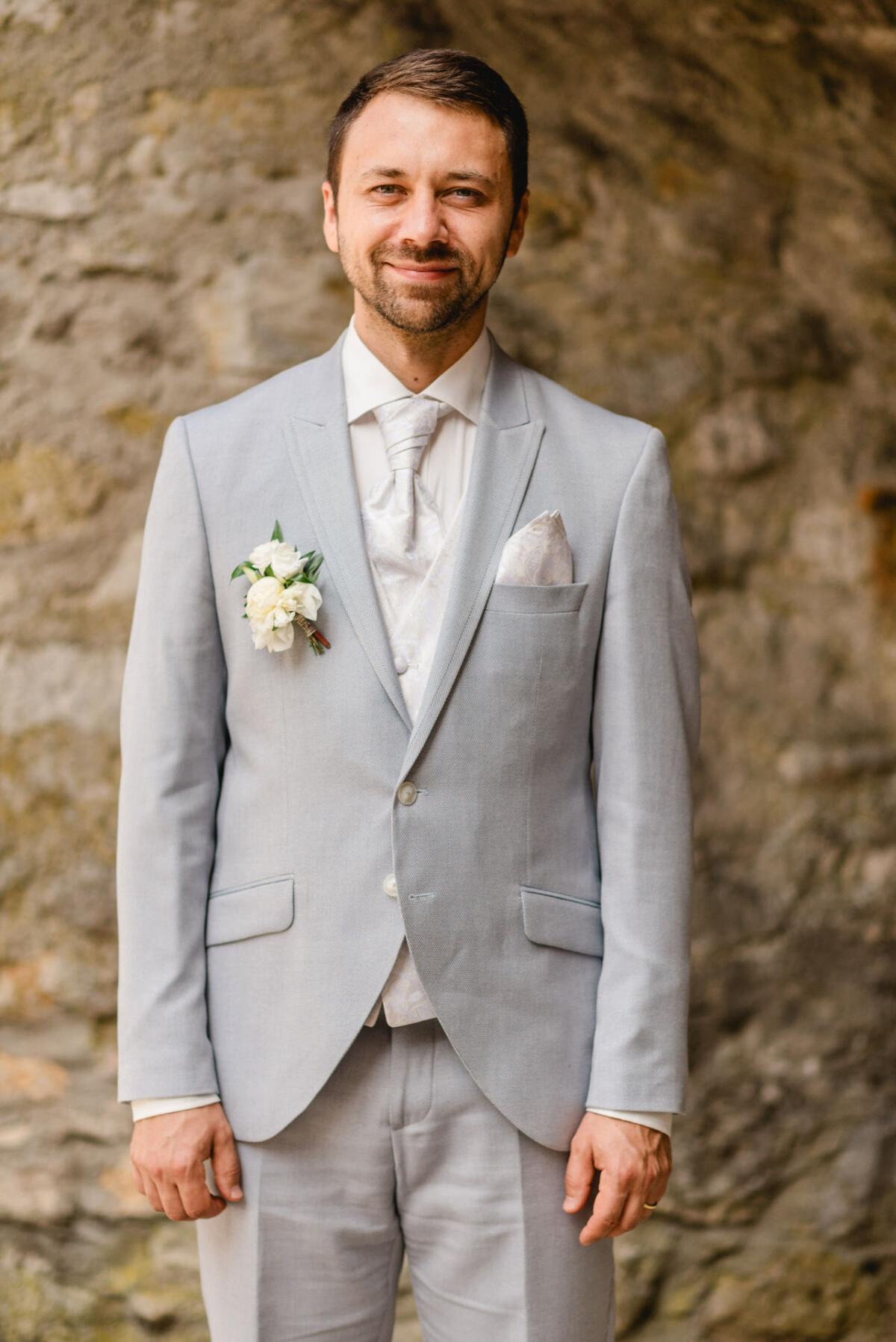 Ein Mann in einem hellgrauen Anzug mit weißem Hemd und Krawatte steht vor einer Steinmauer und trägt eine weiße Ansteckblume.