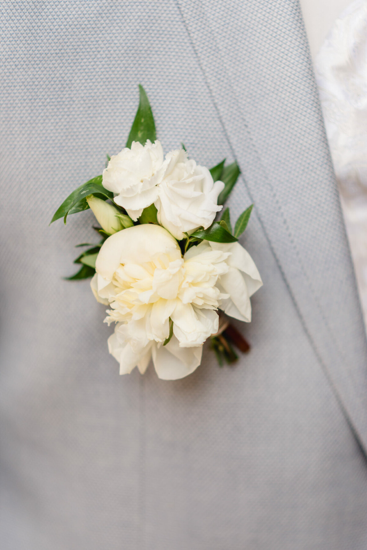 Eine Nahaufnahme einer Boutonniere, die an einem hellgrauen Anzugjackett befestigt ist. Die Boutonniere ist mit weißen Blumen und grünen Blättern verziert.