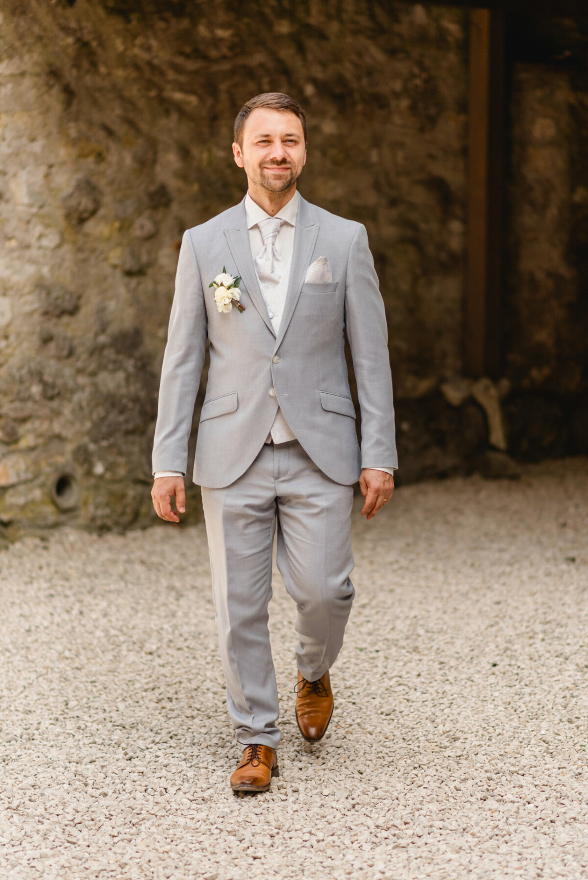 Ein Mann in einem hellgrauen Anzug und braunen Schuhen geht auf einem Kiesweg vor einer Steinmauer.