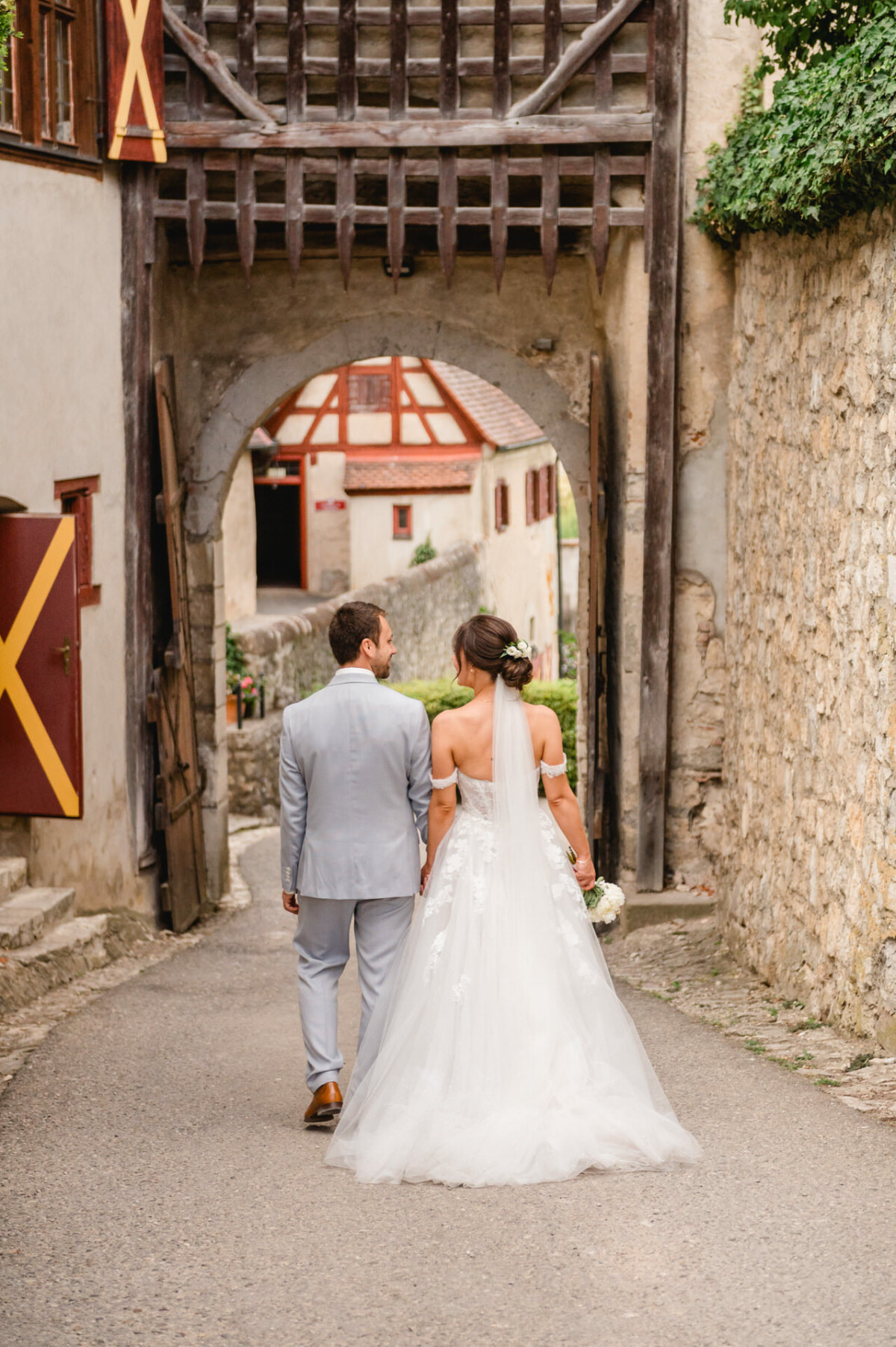 Ein Paar im Hochzeitskleid geht Hand in Hand eine schmale, gepflasterte Gasse unter einem steinernen Torbogen entlang, im Hintergrund sind historische Gebäude zu sehen.