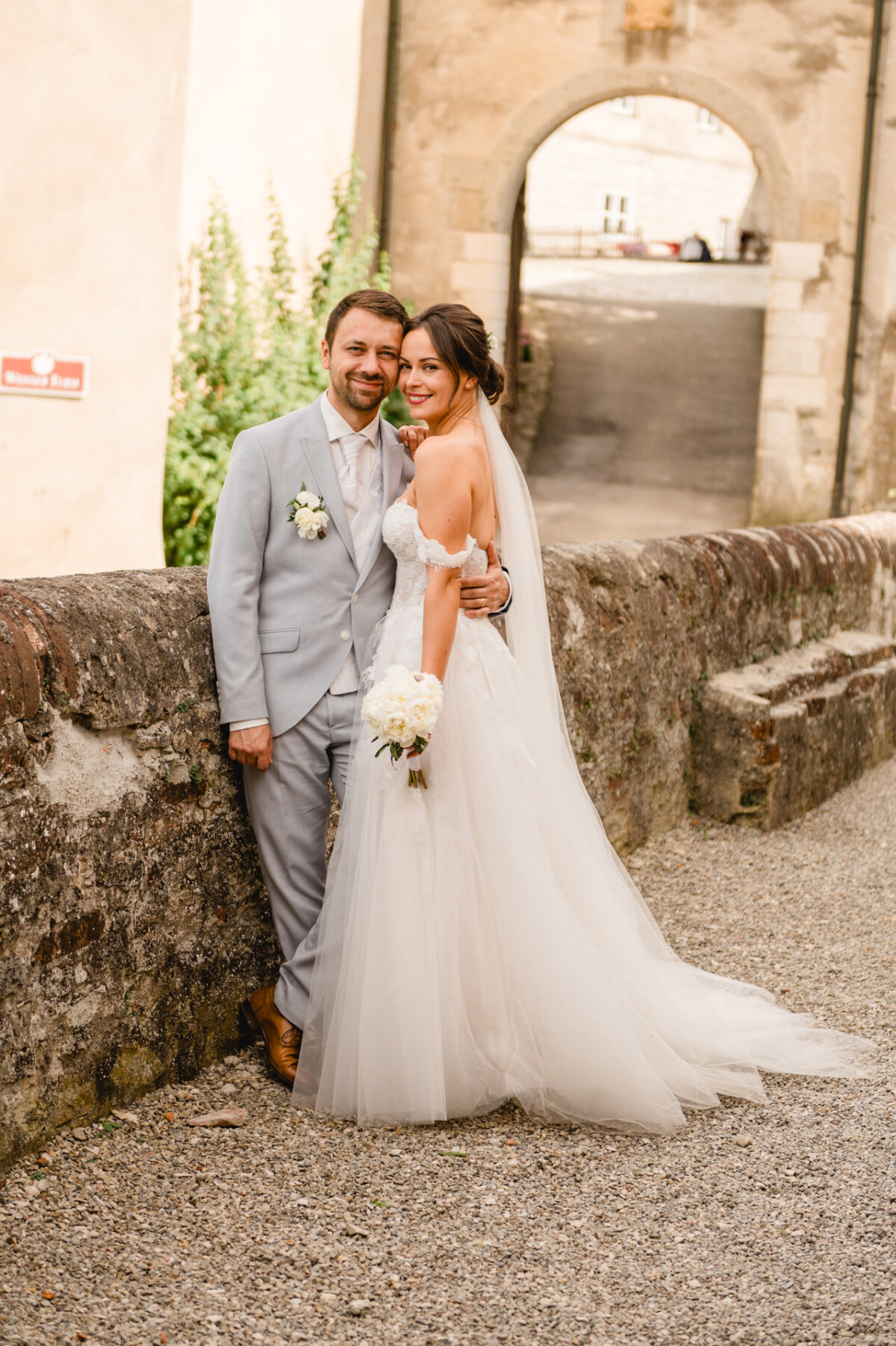 Ein Paar im Hochzeitskleid posiert gemeinsam auf einem Kopfsteinpflasterweg neben einer Steinmauer, im Hintergrund sind ein Gebäude und ein Torbogen zu sehen.
