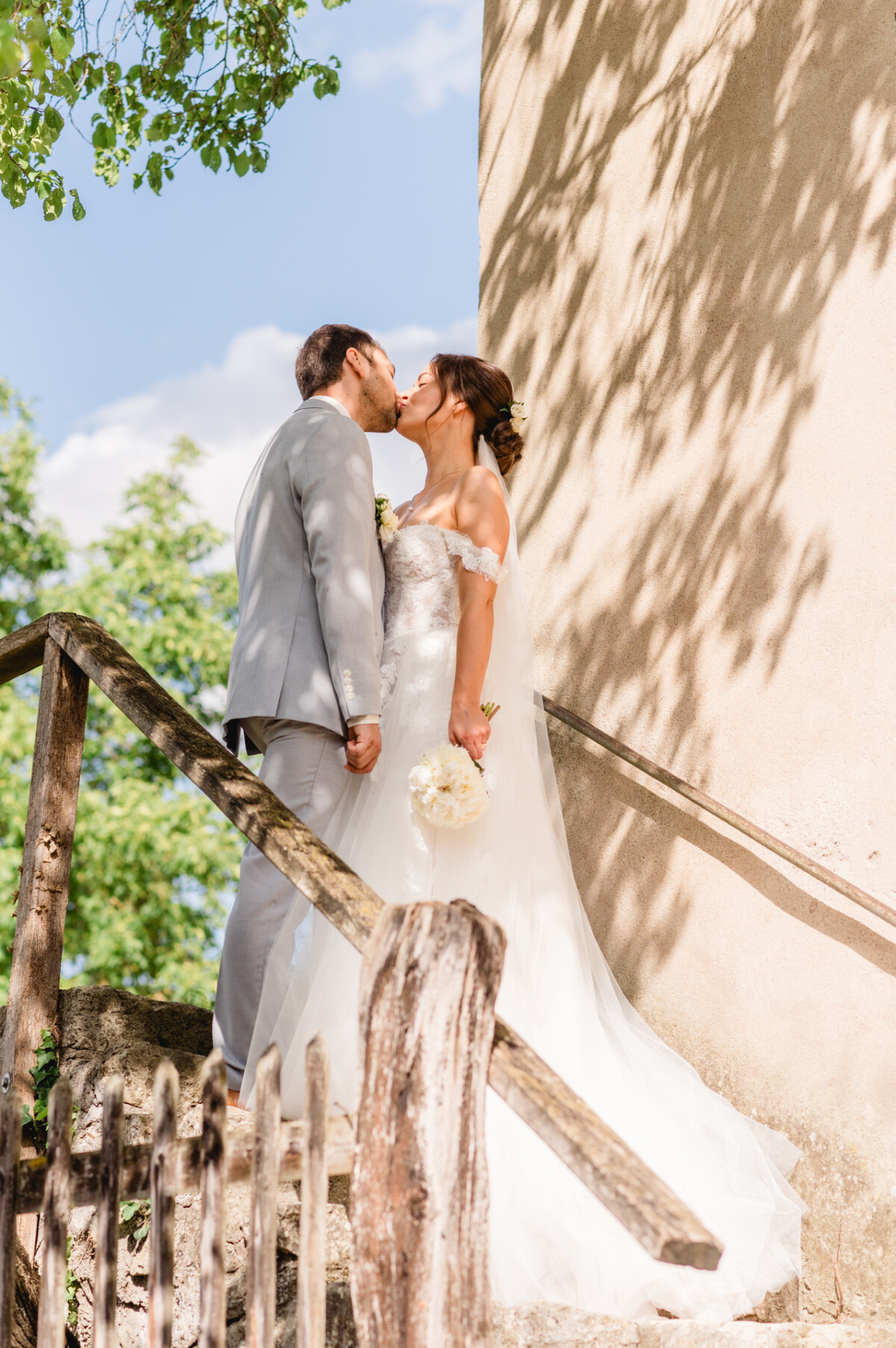Ein Paar im Hochzeitskleid küsst sich auf einer Steintreppe im Freien, im Hintergrund fällt das Sonnenlicht durch die Blätter.