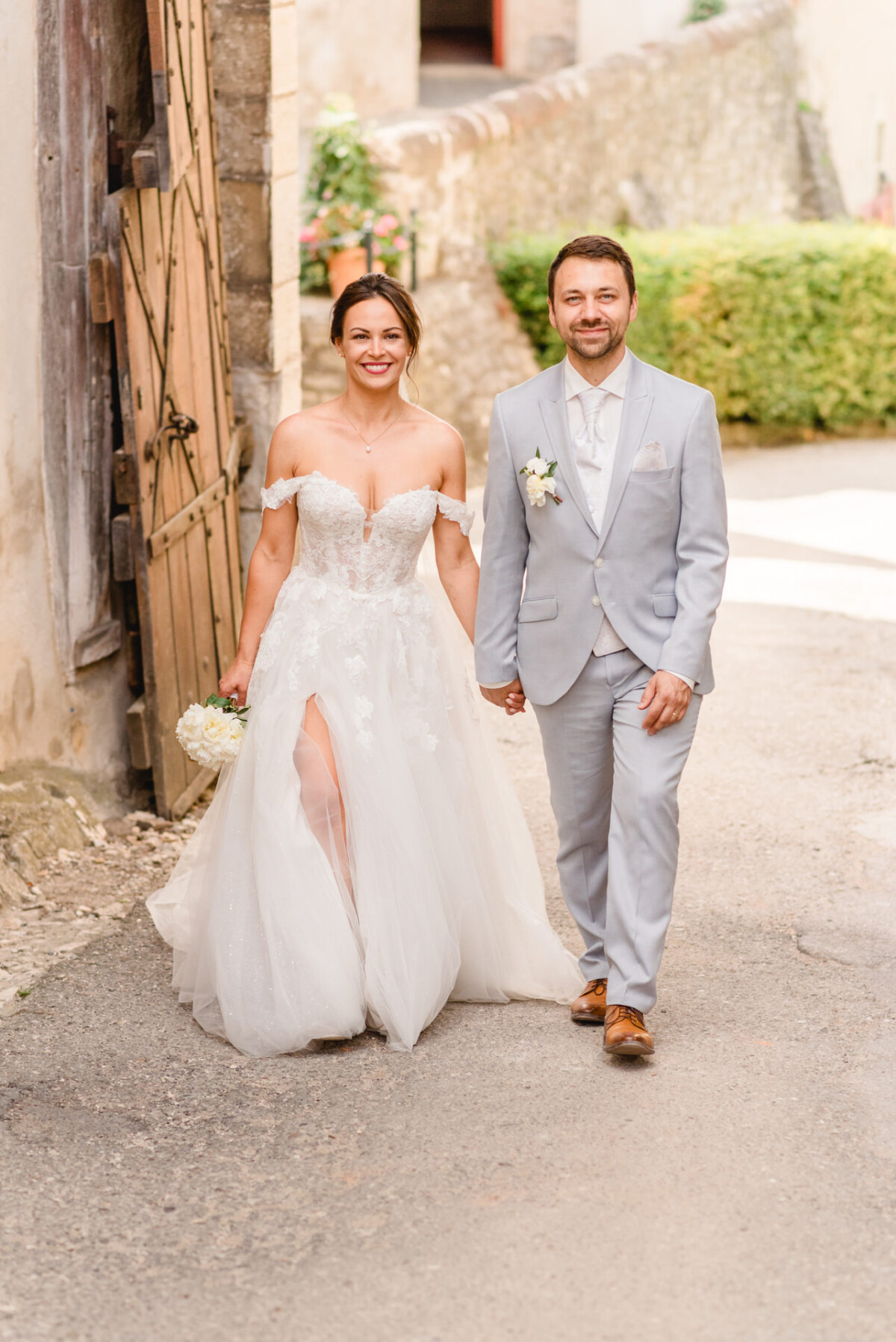 Braut und Bräutigam in Abendgarderobe gehen Hand in Hand auf einer sonnenbeschienenen Straße. Die Braut trägt ein weißes Kleid mit Schlitz und der Bräutigam einen hellgrauen Anzug.
