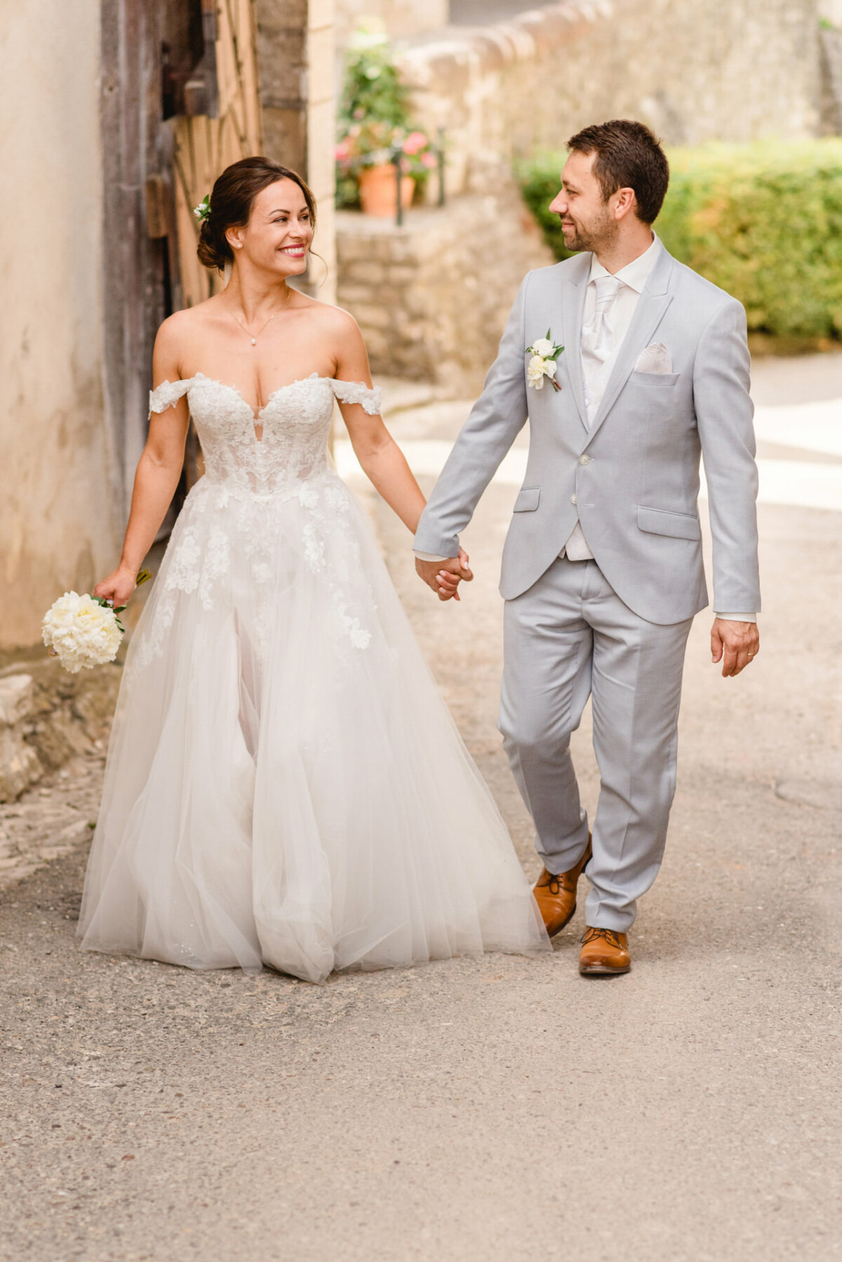 Braut und Bräutigam gehen im Freien an den Händen. Die Braut trägt ein weißes Kleid und hält einen Blumenstrauß, während der Bräutigam einen hellgrauen Anzug trägt.
