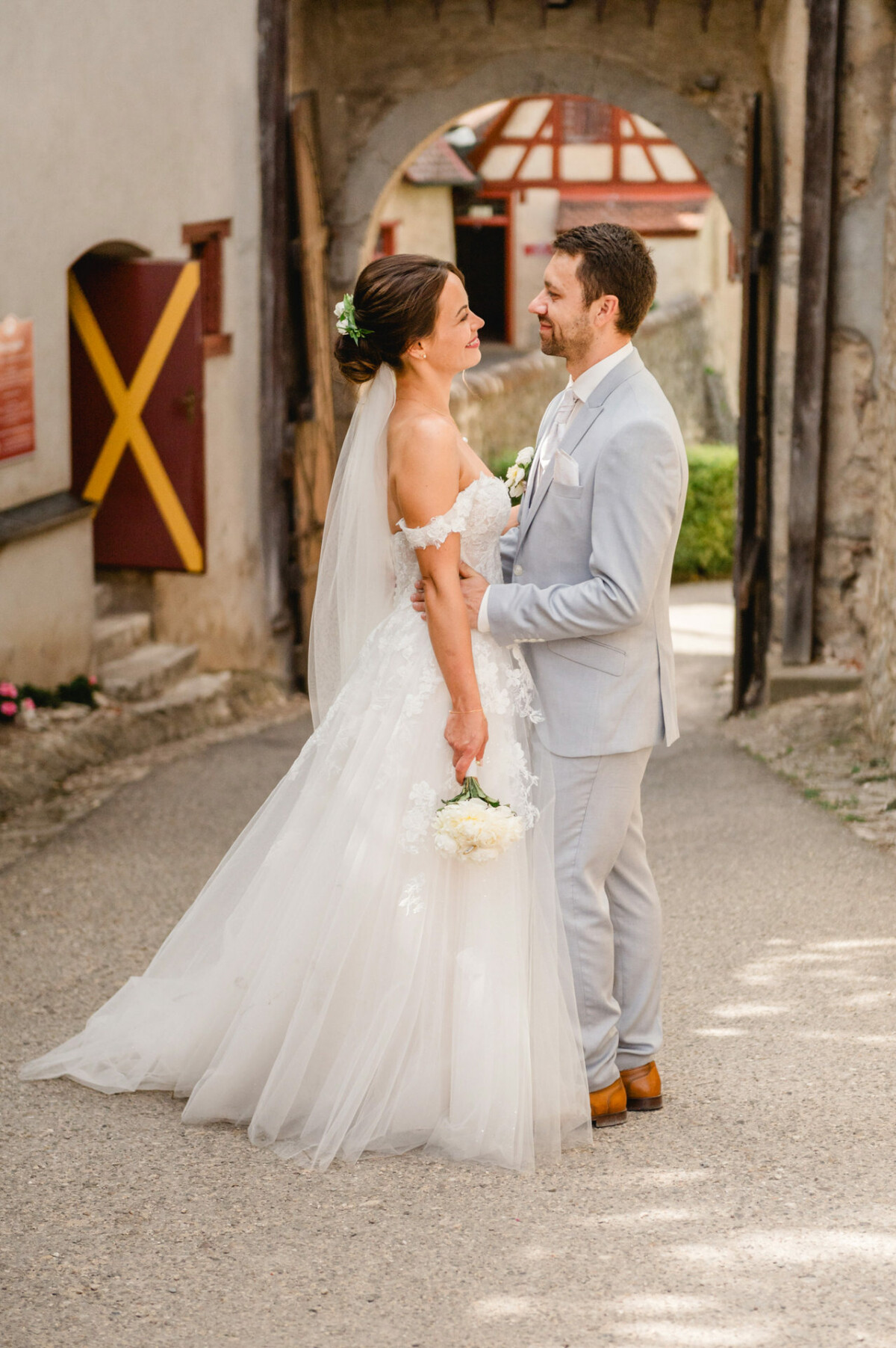 Braut im weißen Kleid und Bräutigam im hellgrauen Anzug stehen einander gegenüber, halten Händchen und lächeln unter einem rustikalen Torbogen.