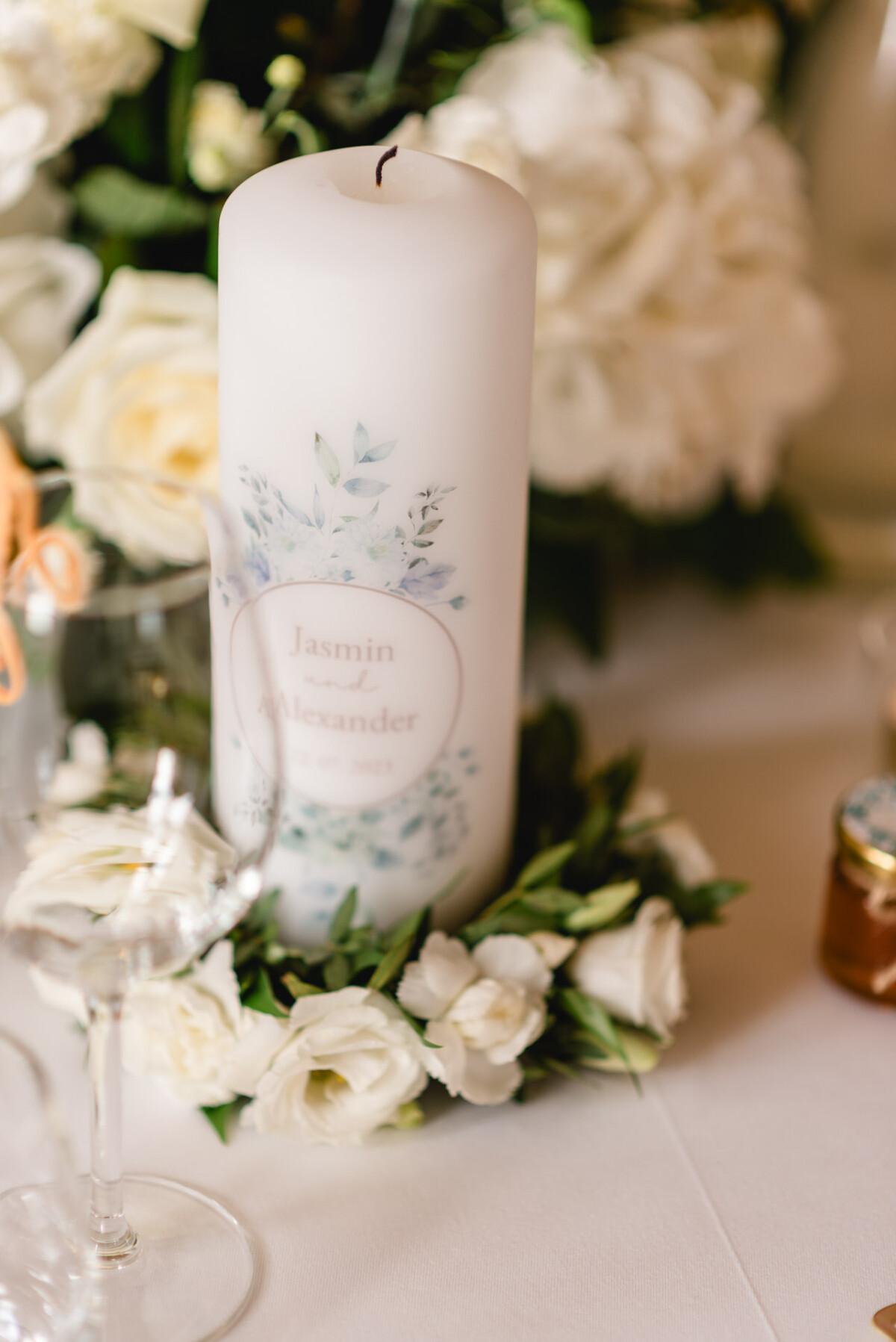 Eine weiß dekorierte Kerze steht auf einem Tisch, umgeben von weißen Blumen, neben einem Weinglas und einem kleinen Gefäß.