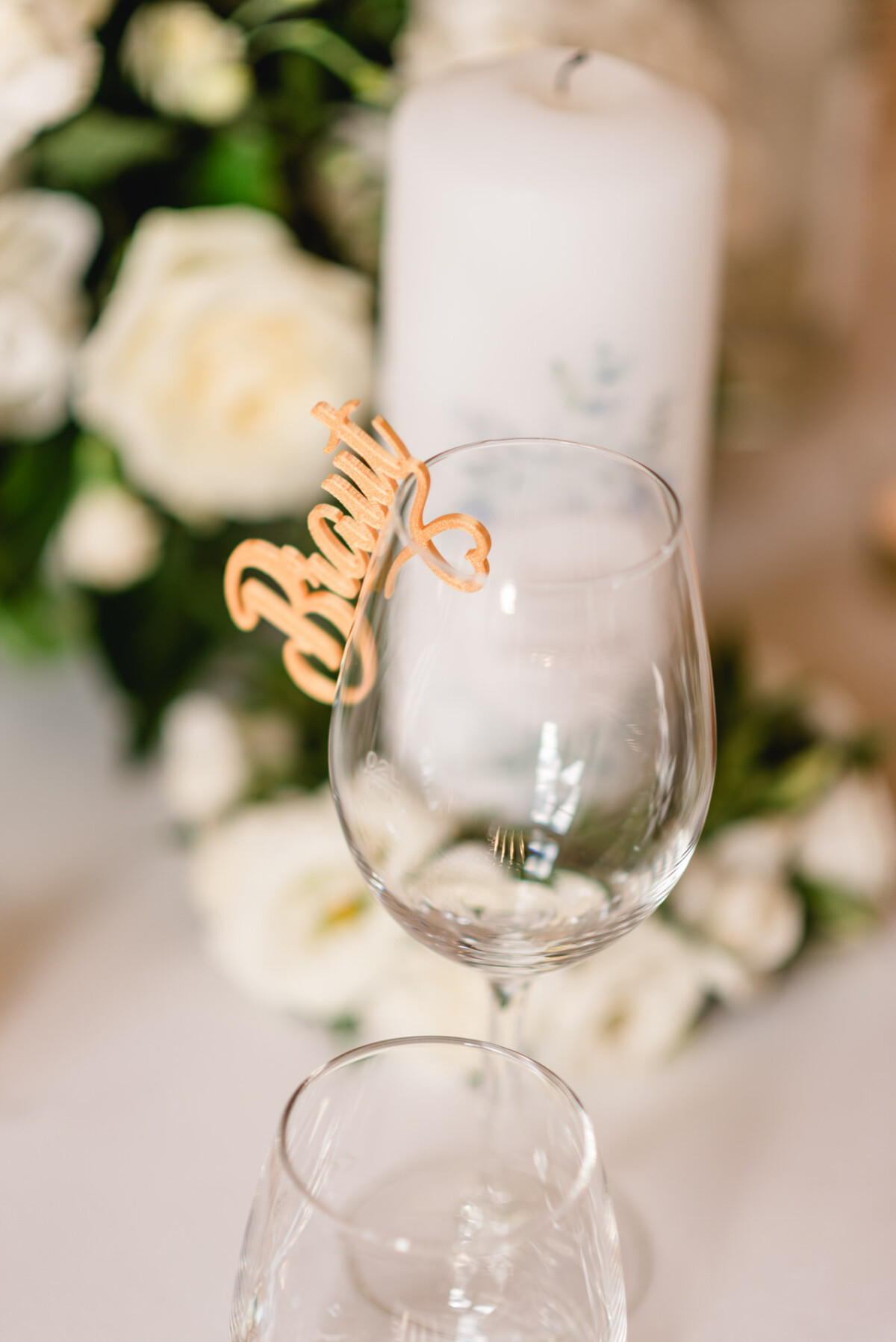 Auf einem Tisch steht ein Weinglas mit dem Etikett „Brandy“ in Schreibschrift, umgeben von weißen Blumen und einer weißen Kerze im Hintergrund.