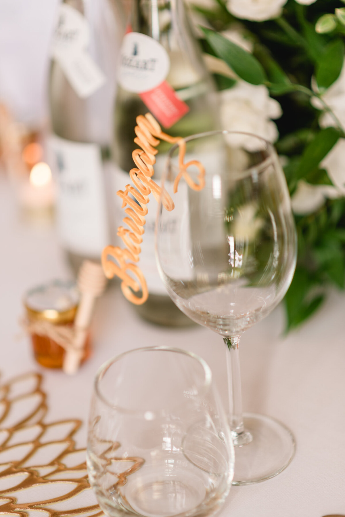 Zwei Weingläser auf einem Tisch, eines mit einem "Bräutigam"-Schild. Flaschen und ein kleines Glas Honig daneben. Weiße Blumen im Hintergrund.