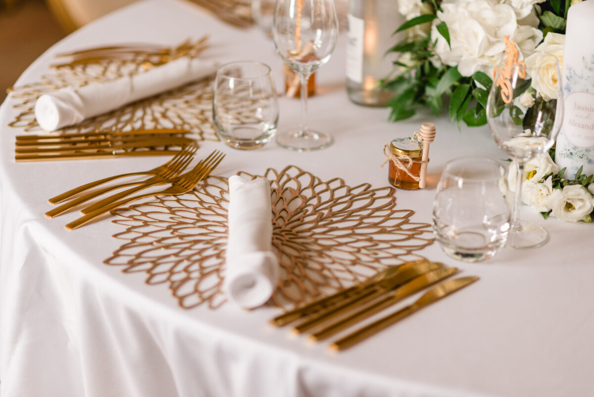 Elegante Tischdekoration mit weißer Tischdecke, goldenem Besteck und Tischsets mit Blumenmotiv. Gerollte Servietten und Glaswaren stehen neben dekorativen Blumen und einem kleinen Honigglas als Tafelaufsatz.