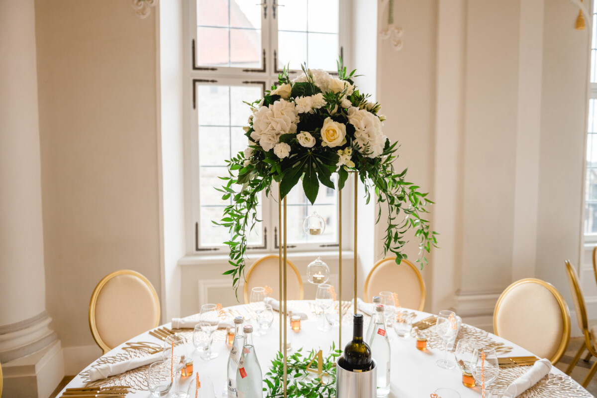 Hohes Blumengesteck mit weißen Rosen und Grünpflanzen auf einem runden Tisch für eine Veranstaltung mit goldenen Stühlen, Wasserflaschen und Glaswaren in einem hellen Raum mit großen Fenstern.