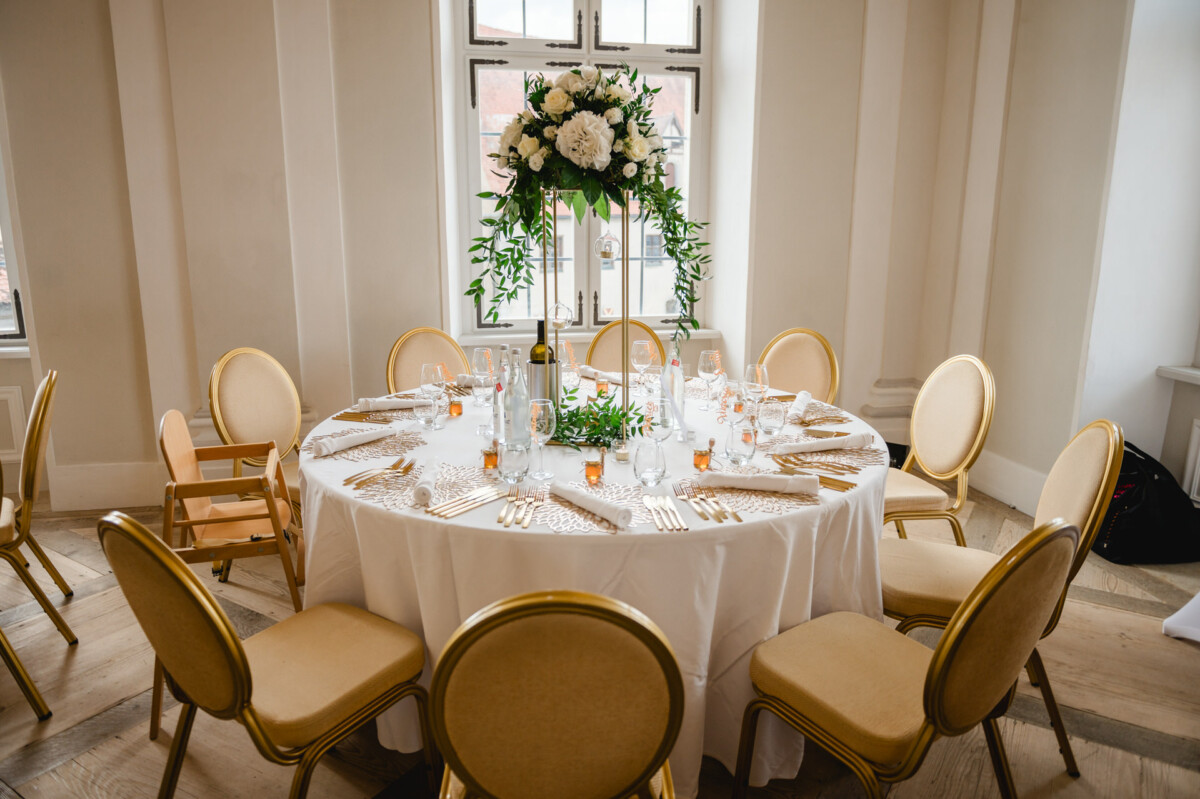 Runder Tisch für eine formelle Veranstaltung mit weißer Tischdecke, goldenen Stühlen, Blumenschmuck, Glaswaren und Besteck.