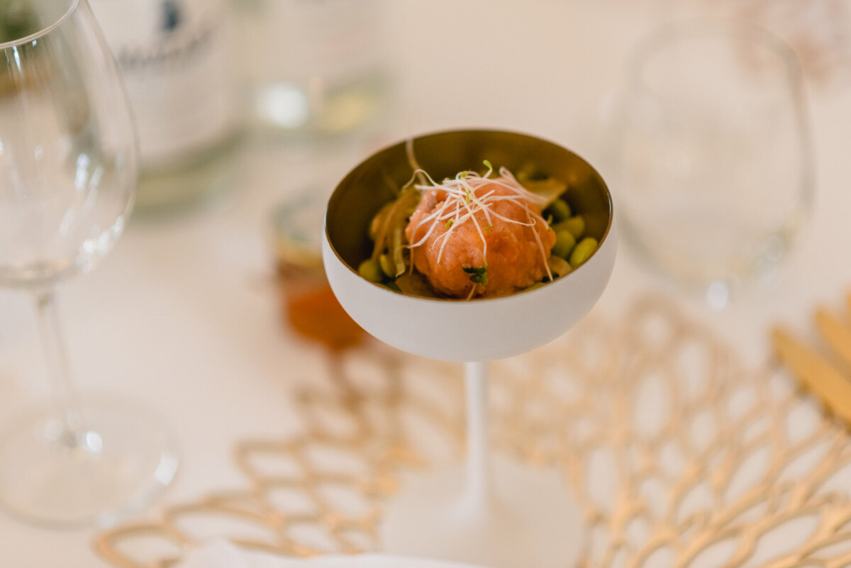 Eine kleine Vorspeise mit runder Portion, garniert mit Sprossen, serviert in einer flachen Schale auf einem Stiel, platziert auf einem geflochtenen Tischset. Im Hintergrund leere Weingläser und Wasserflaschen.
