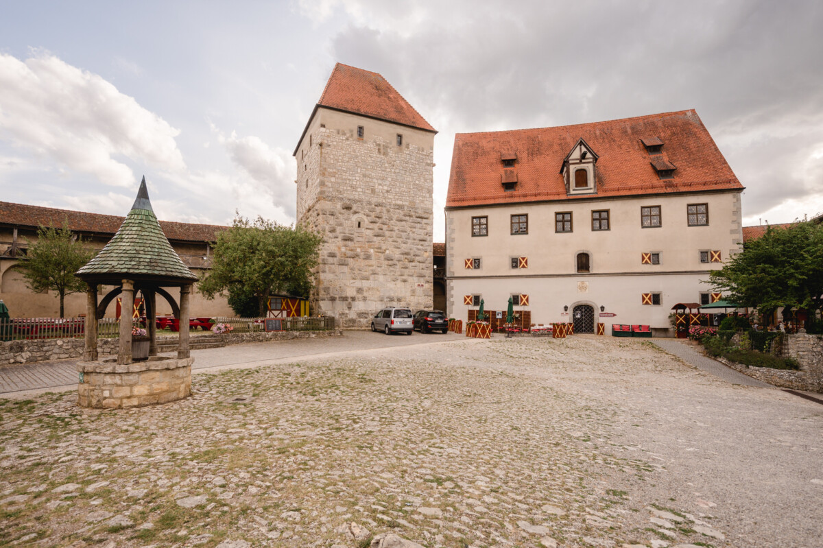 Historischer Schlosshof mit einem Steinturm, einem Gebäude mit roten Dächern und einem Brunnen. Unter einem teilweise bewölkten Himmel sind mehrere geparkte Autos auf einer Kopfsteinpflasterfläche zu sehen.