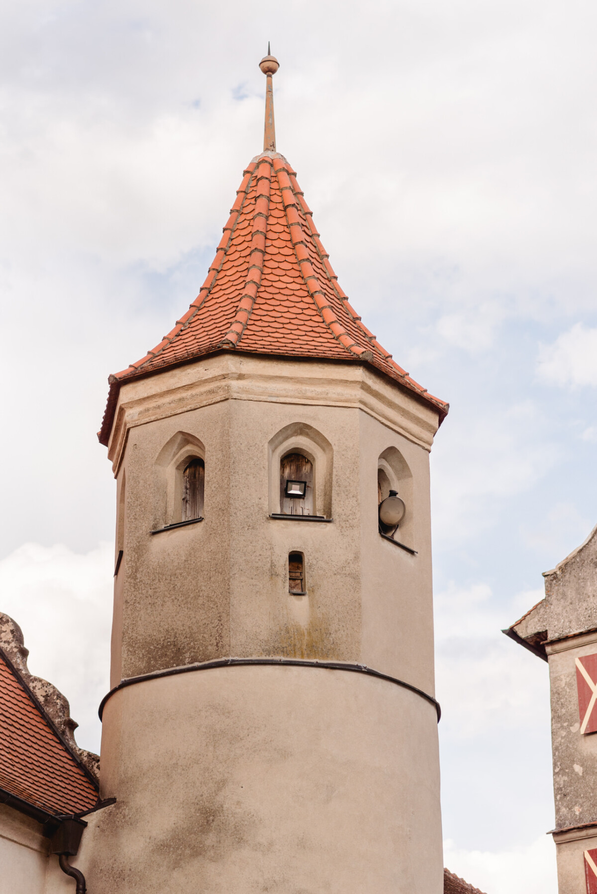 Ein Steinturm mit einem kegelförmigen Dach aus roten Ziegeln und kleinen Bogenfenstern vor einem bewölkten Himmel.