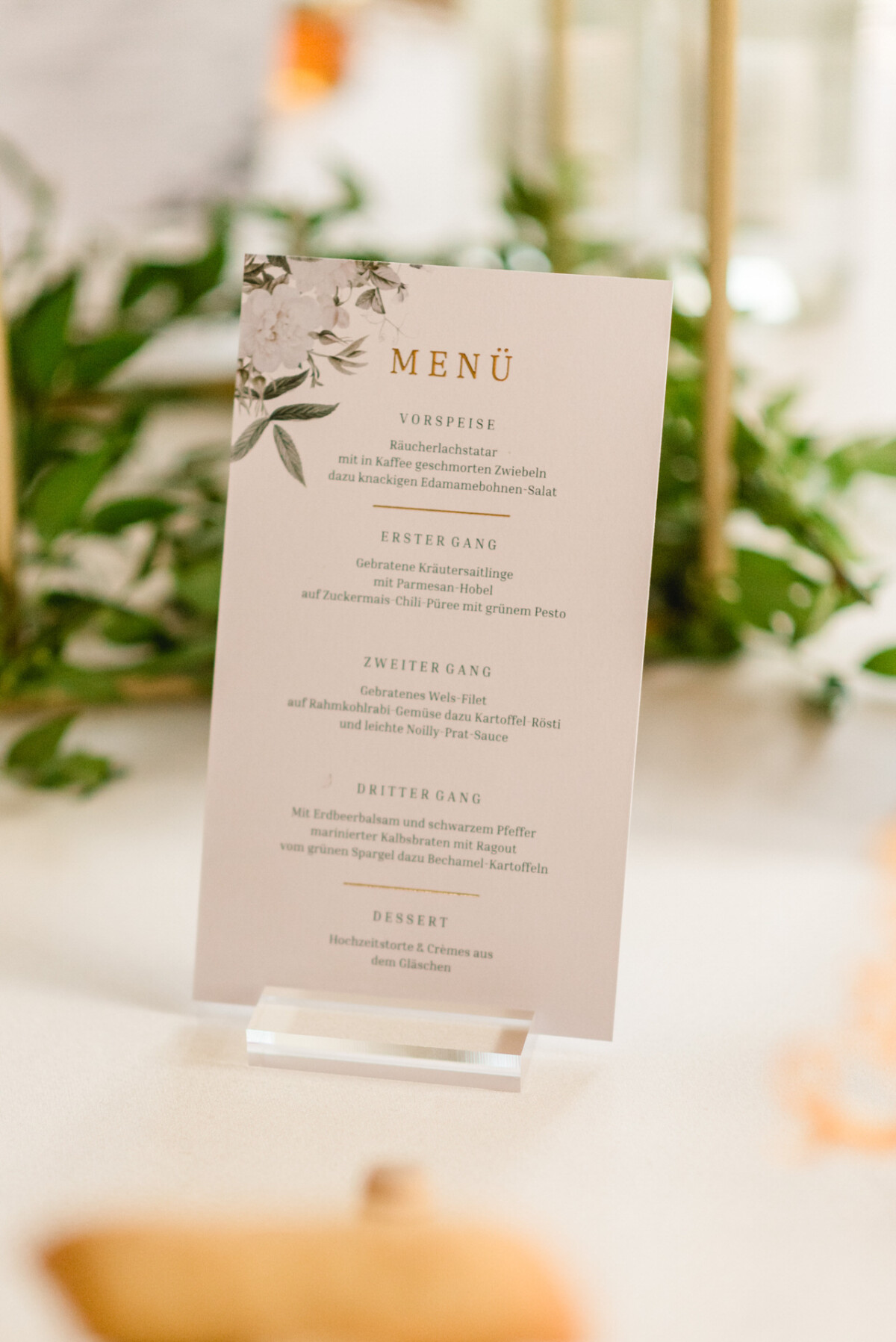 Eine gedruckte Menükarte auf einem Ständer mit floralem Design, auf der eine Auswahl an Gängen in deutscher Sprache aufgeführt ist.