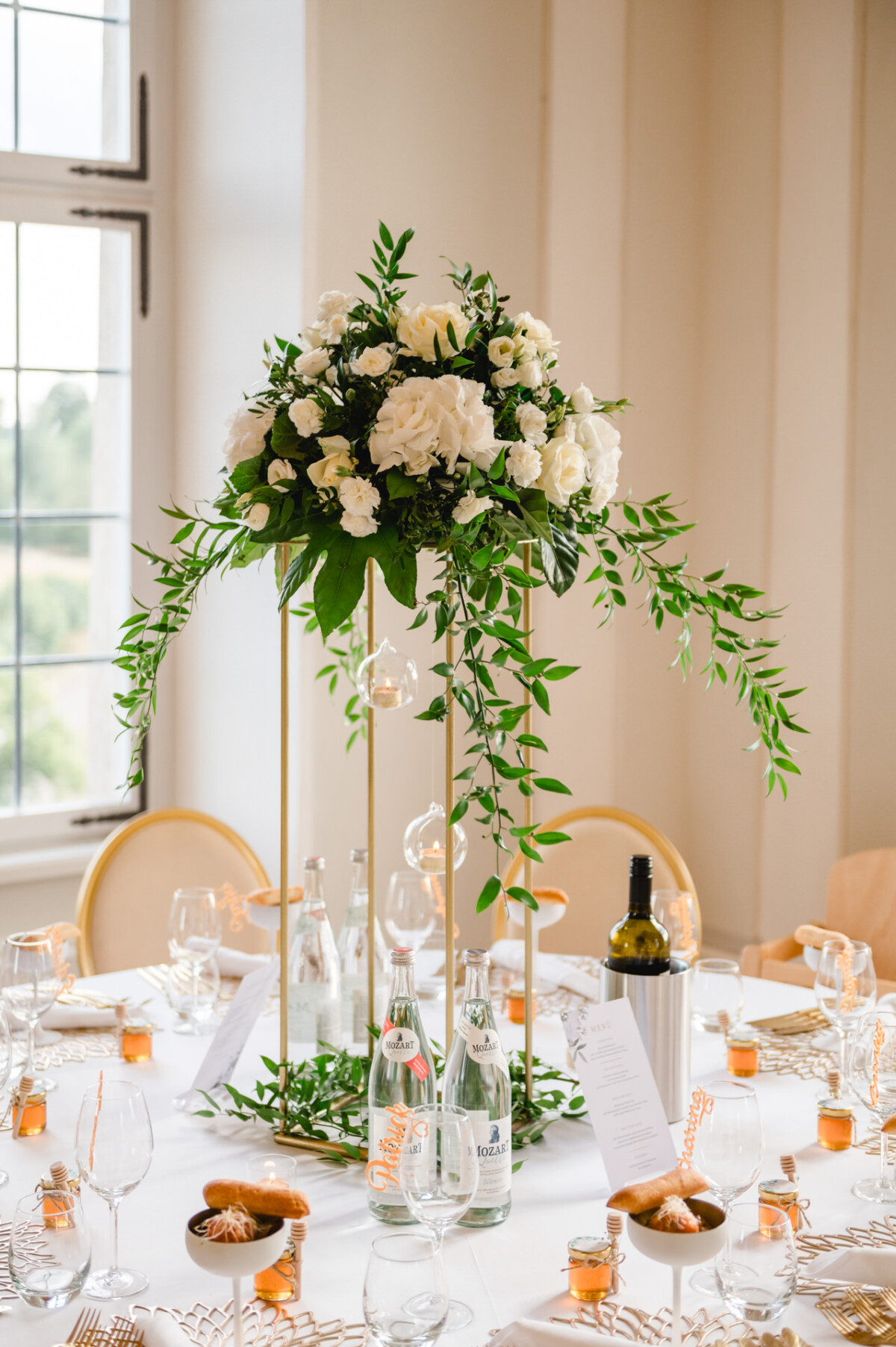 Elegante Tischdekoration mit einem hohen Blumengesteck mit weißen Blumen und Grünpflanzen, umgeben von Weingläsern, Wasserflaschen, kleinen Orangengläsern und einer Weinflasche.