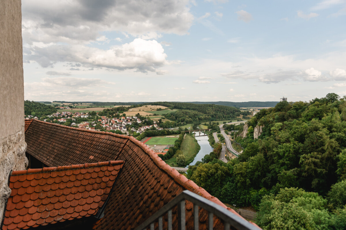 Malerische Aussicht von einem Dach auf einen Fluss, bewaldete Hügel und eine kleine Stadt unter einem bewölkten Himmel.