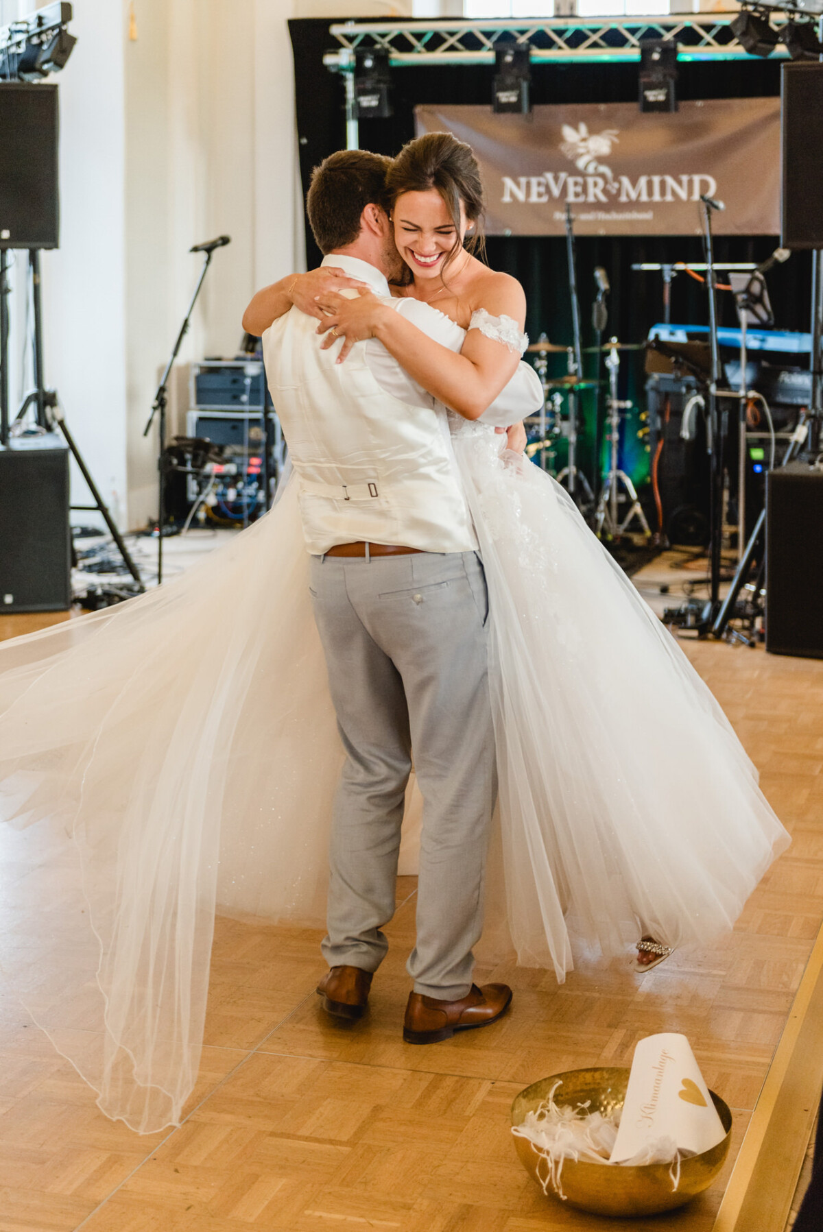 Braut und Bräutigam umarmen sich auf einer Tanzfläche. Im Hintergrund ist ein Bandaufbau zu sehen und im Vordergrund steht eine dekorierte Schüssel.