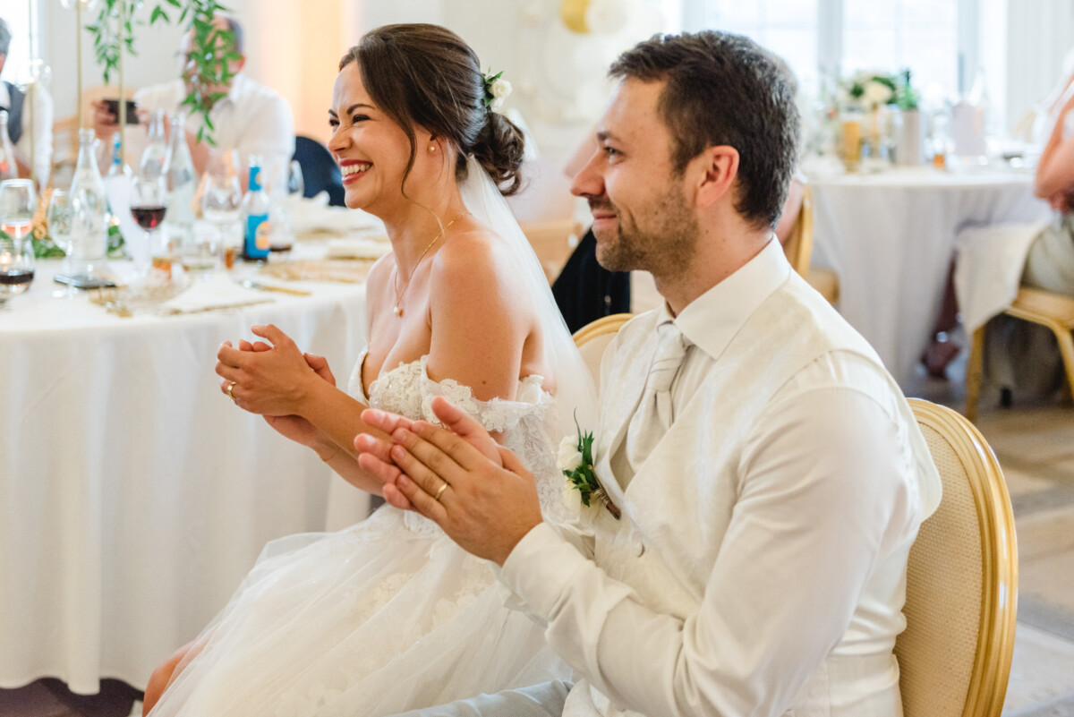Eine Braut und ein Bräutigam sitzen während eines Hochzeitsempfangs an einem Tisch, lächeln und klatschen.