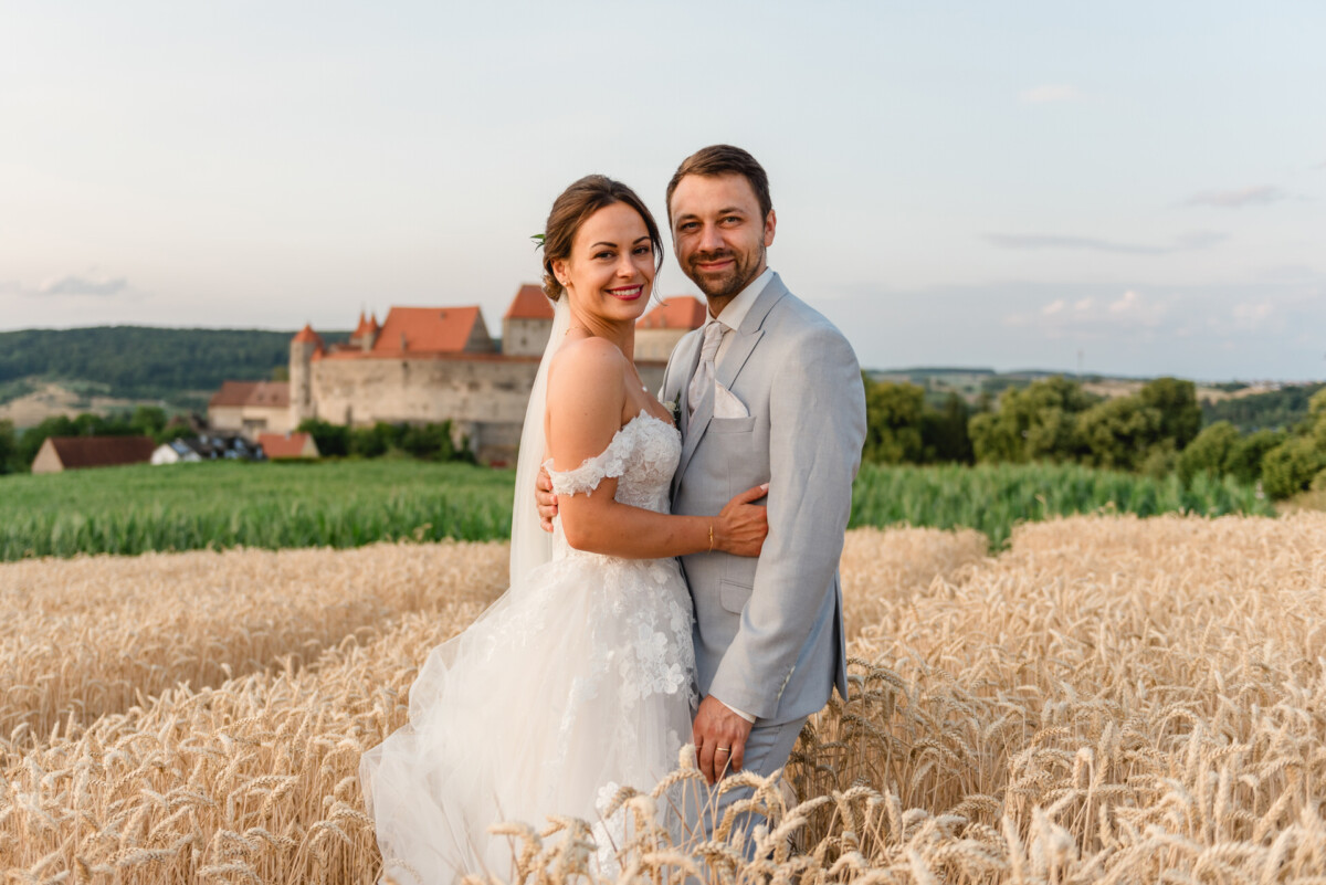 Eine Braut und ein Bräutigam stehen lächelnd in einem Weizenfeld, im Hintergrund ist ein Schloss zu sehen.