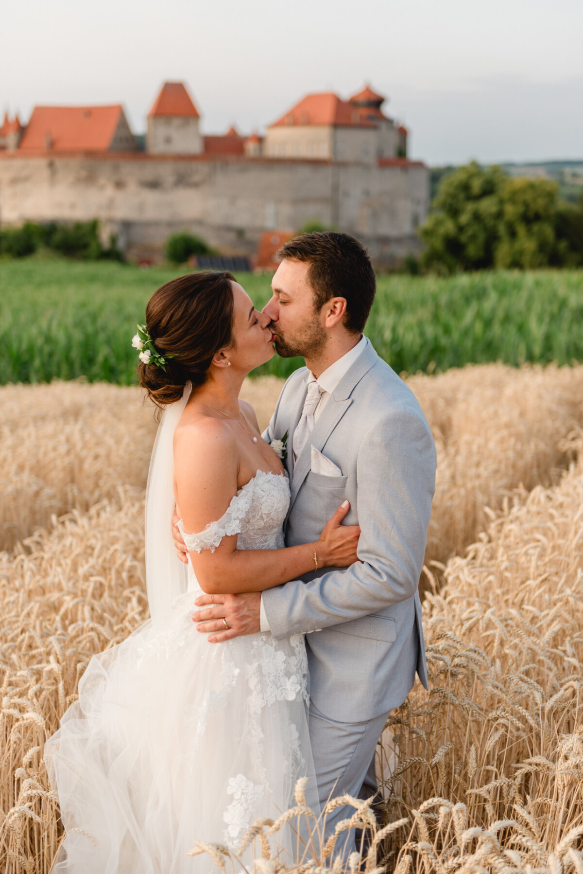 Braut und Bräutigam küssen sich in einem Weizenfeld mit einer mittelalterlichen Burg im Hintergrund.
