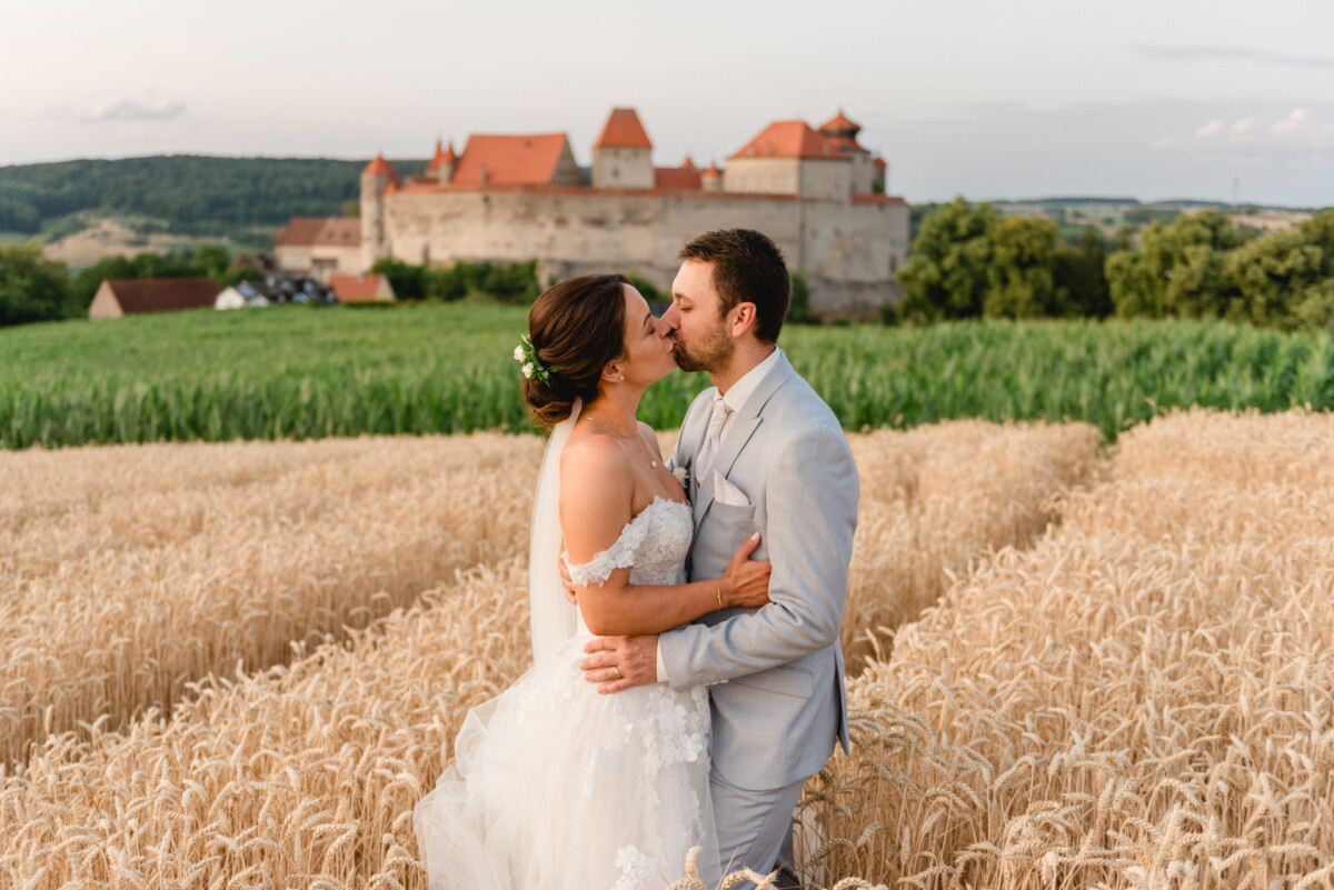 Braut und Bräutigam küssen sich in einem Weizenfeld mit einem Schloss im Hintergrund.