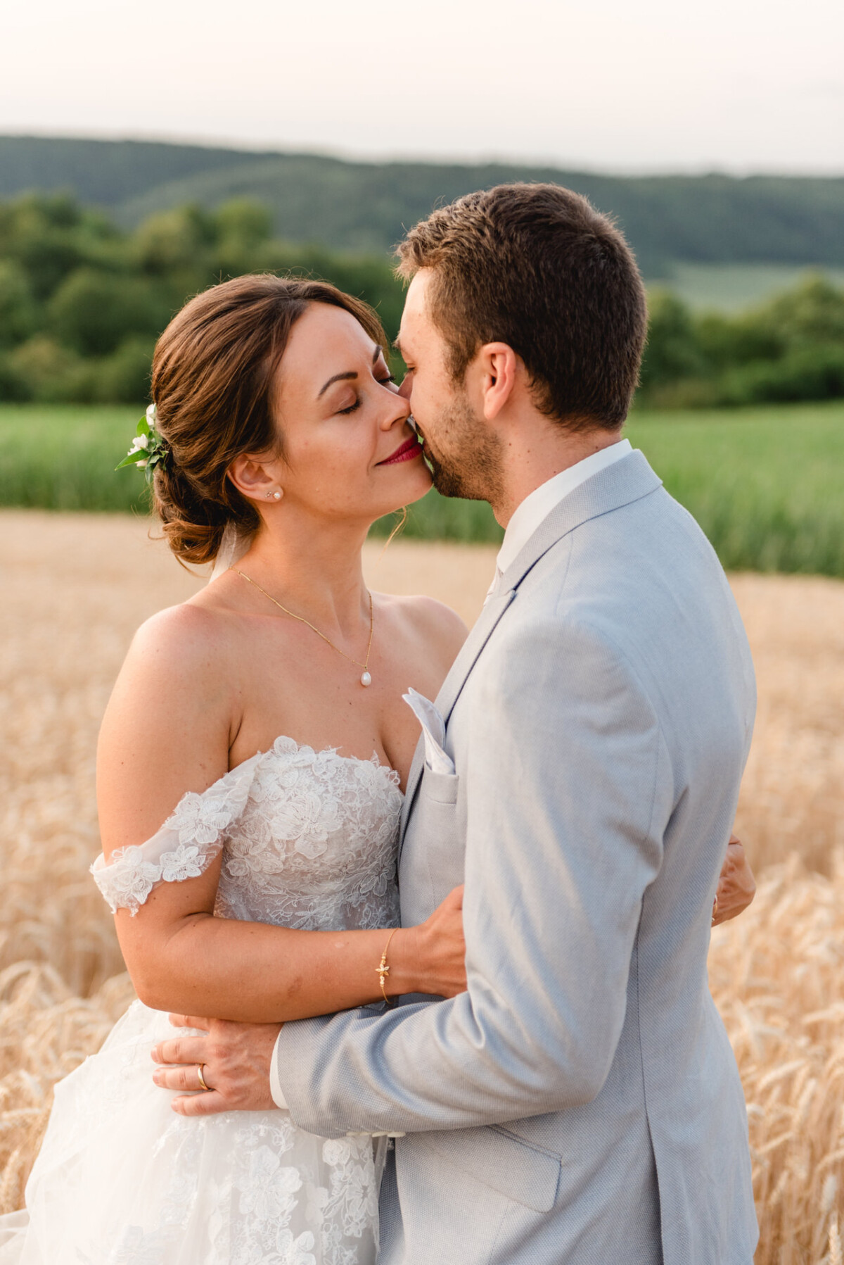 Ein Paar im Hochzeitskleid umarmt und küsst sich auf einem Feld vor der Kulisse sanfter Hügel und grüner Landschaft.