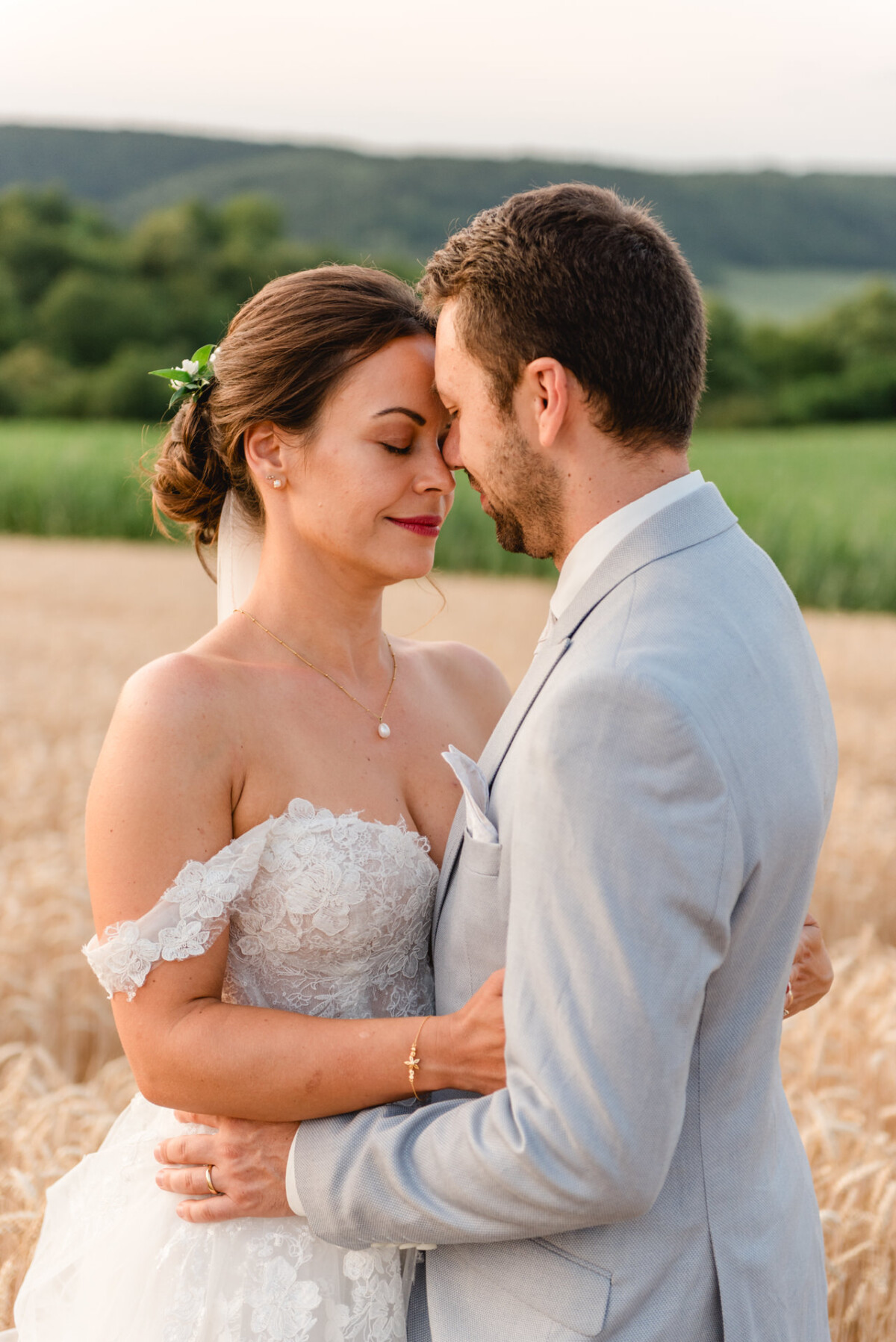 Braut und Bräutigam umarmen sich in einem Weizenfeld und berühren sanft ihre Stirnen. Die Braut trägt ein schulterfreies Spitzenkleid und der Bräutigam einen hellgrauen Anzug. Im Hintergrund sind grüne Hügel zu sehen.