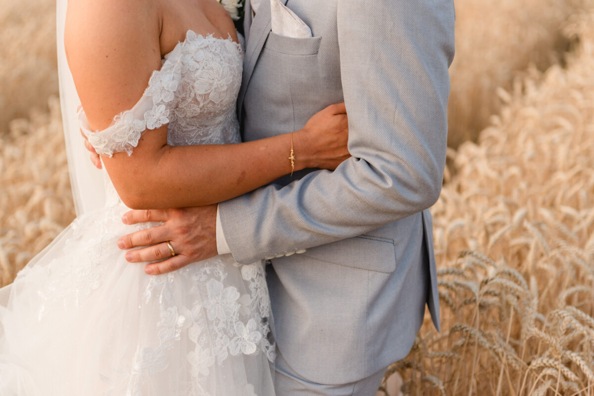 Ein Paar im Hochzeitskleid umarmt sich in einem Weizenfeld. Die Braut trägt ein Spitzenkleid und der Bräutigam einen hellgrauen Anzug.